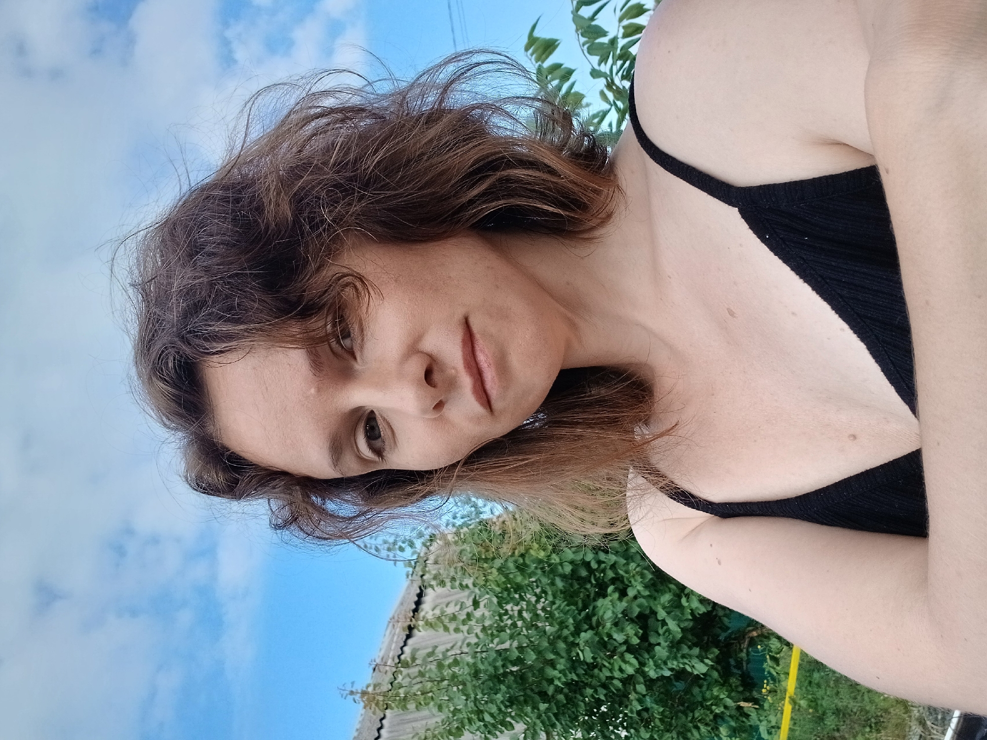 A woman with shoulder-length brown hair is pictured in a selfie taken outdoors. She is wearing a black tank top and has her arms crossed. Behind her, a green tree and a grey, corrugated metal roof are visible against a blue sky with scattered clouds. The environment appears to be rural, possibly in Maslova Pristan', Russia, with the presence of a residential building and vegetation.
