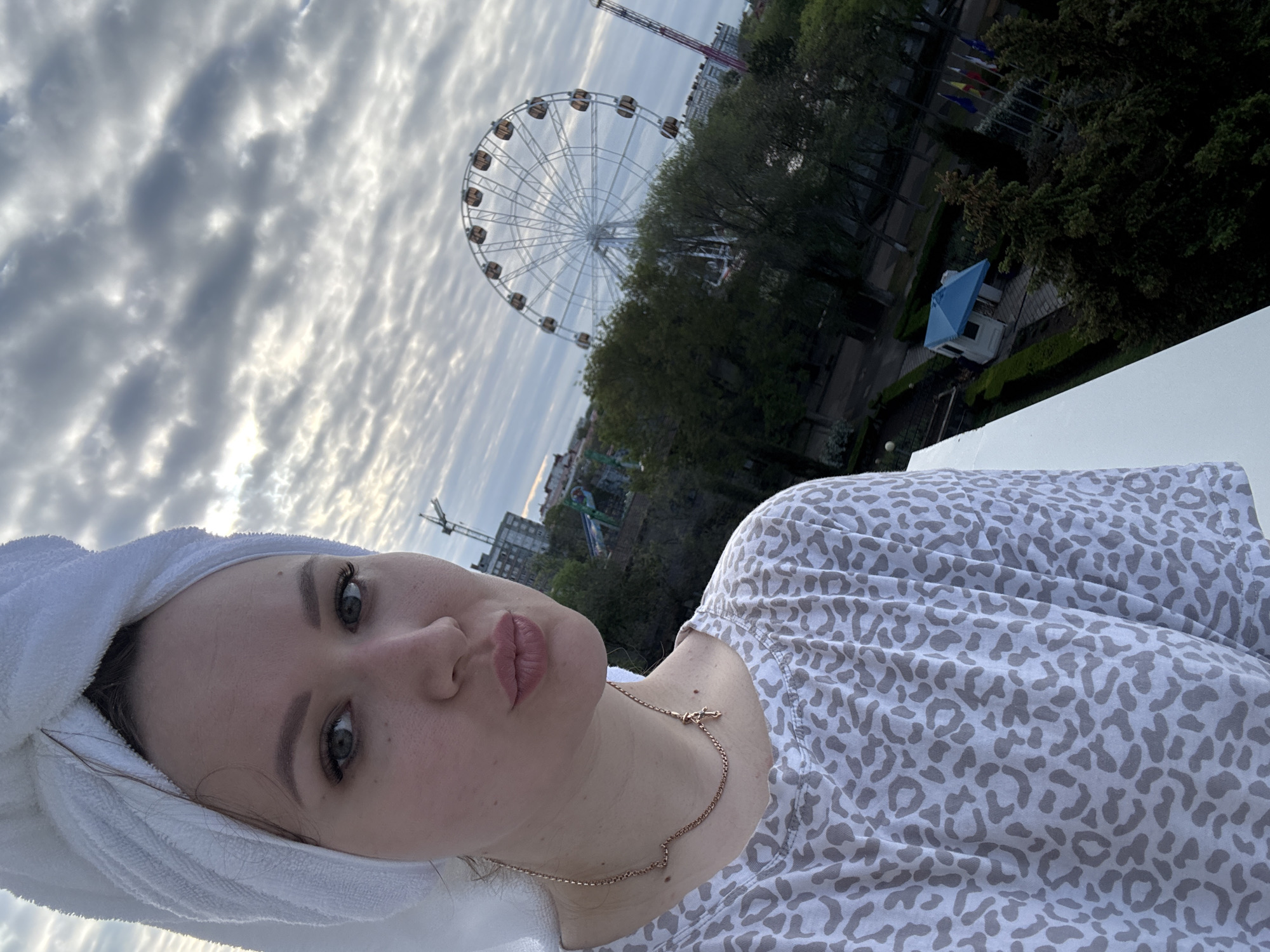 A young woman, wearing a white towel wrapped around her head and a light gray patterned shirt, poses in a selfie with a cityscape visible in the background. Her lips are pursed. A gold necklace is visible around her neck. Beyond the white railing she appears to be standing behind, a ferris wheel rises above the trees, indicating an amusement park. Other structures, including a small blue and white building, additional rides, and a tall crane, are also visible in the background. Flags are also visible. The sky is cloudy. Based on the visual context, specifically the ferris wheel and other background elements, the location is identified as Yevpatoriya, Ukraine.