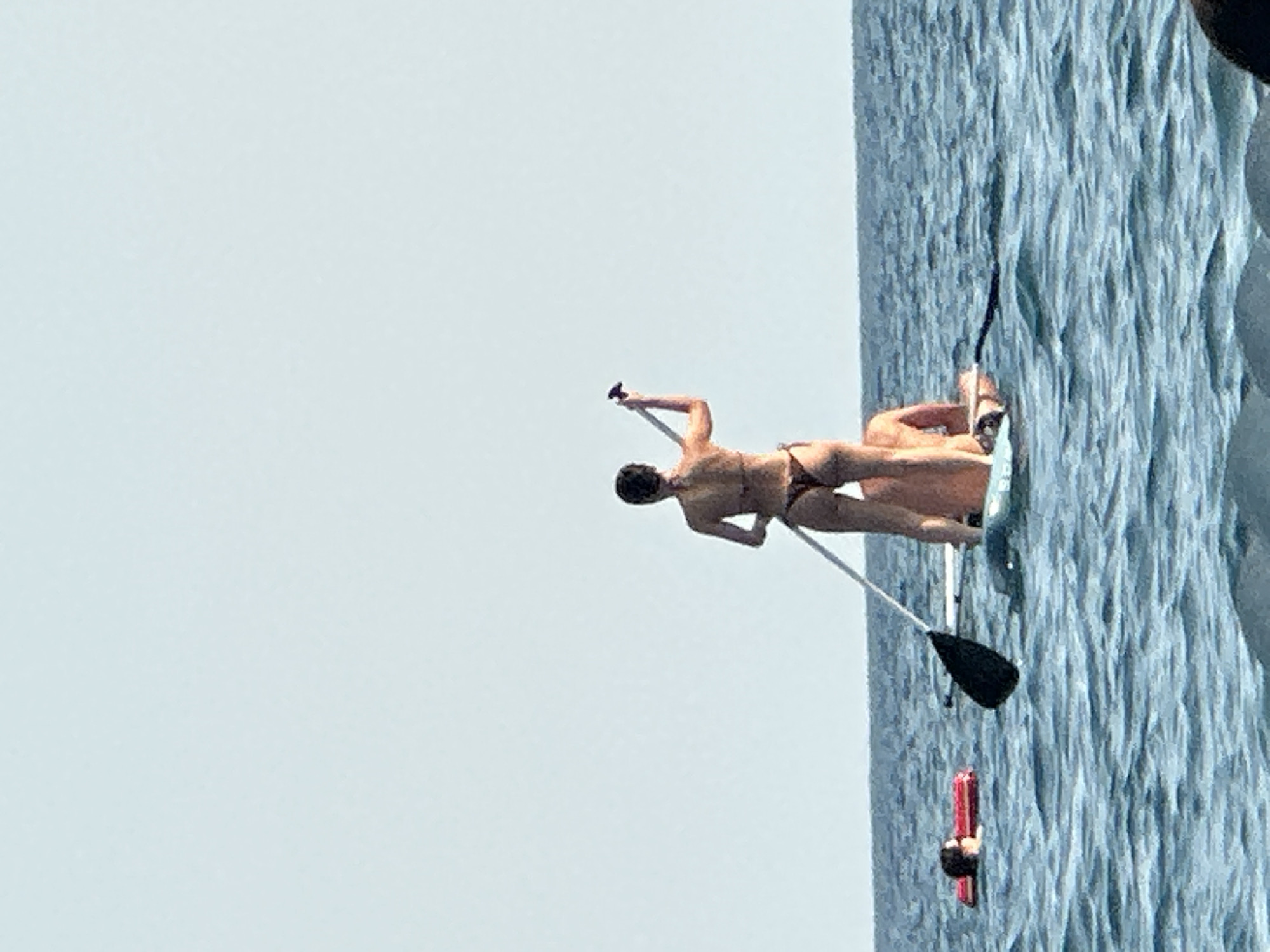 The image depicts a clear sky over a blue sea. In the foreground, two individuals are on a paddleboard. One individual, wearing a bikini, is standing and holding a paddle. Another individual is seated on the paddleboard behind the standing person. To the left of the paddleboard, a person wearing sunglasses is partially submerged in the water, resting on a red inflatable object. The scene suggests a recreational activity on the water. The location is identified as Boreti, Montenegro.
