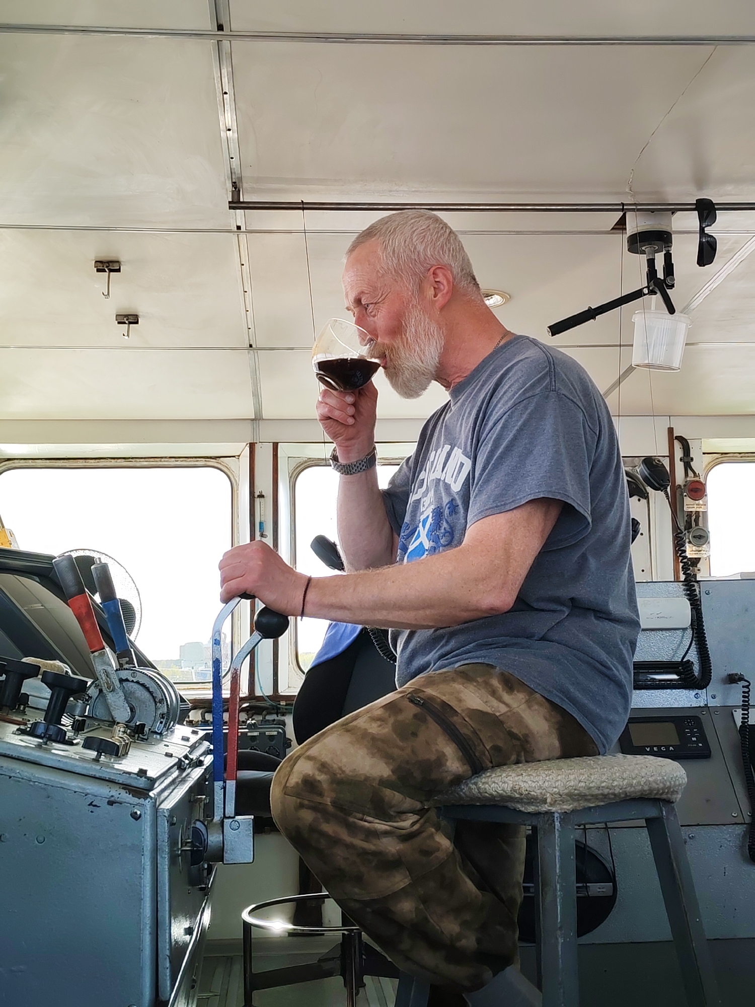The image depicts a man with grey hair and a beard, sitting on a stool in what appears to be the control room of a boat or ship. He is wearing a grey t-shirt and camouflage pants. He is holding a wine glass in his right hand and appears to be drinking from it, while his left hand is holding onto a lever. The control panel in front of him is a metallic grey with various levers, switches, and gauges. There is a window behind him, and the outside scene shows what looks like land or possibly another ship. The interior of the room is utilitarian with a metal ceiling and white walls. The location cannot be determined from the image.