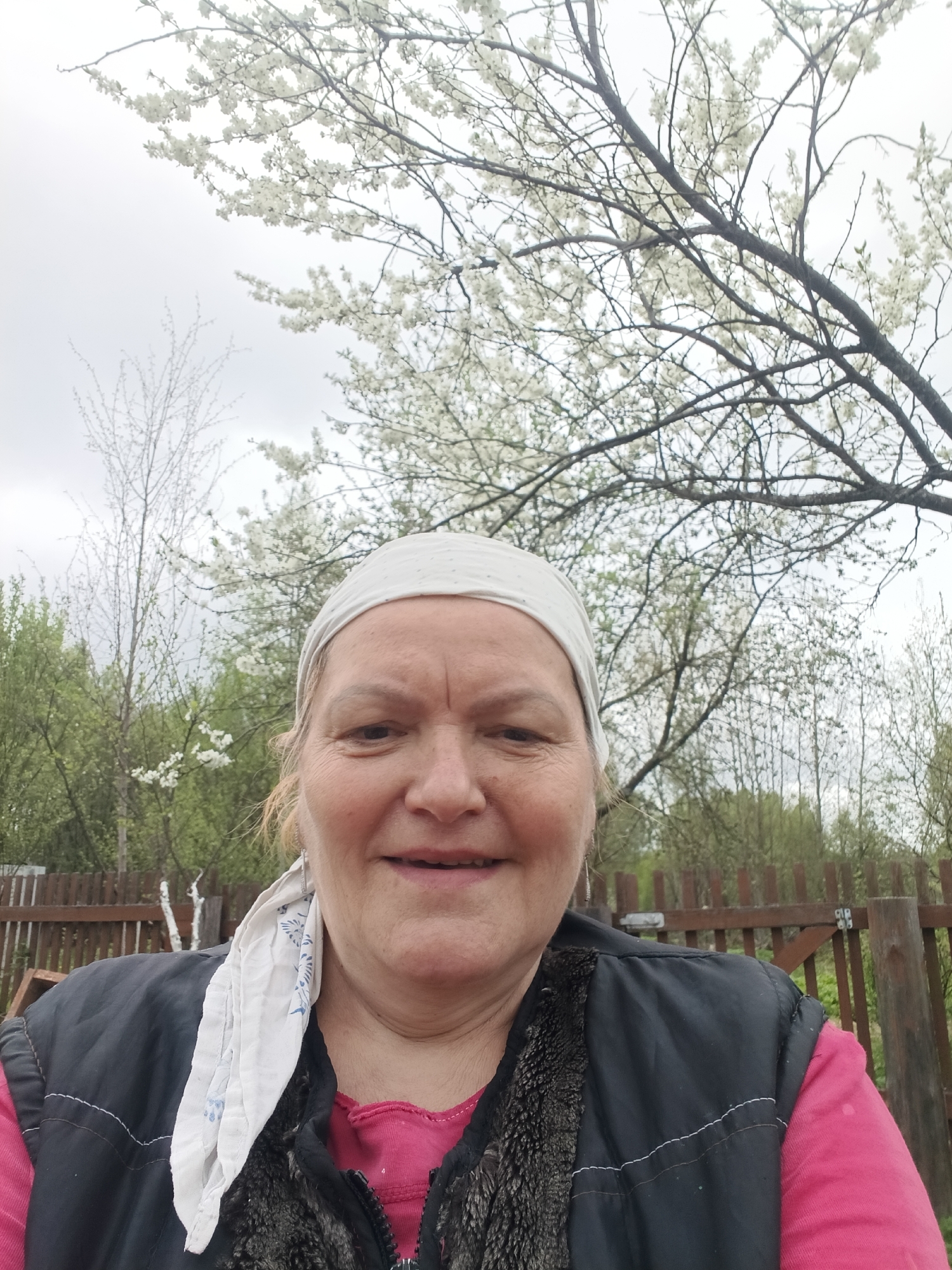 An adult woman stands outside. She wears a pink shirt, a black vest with a fur collar, and a white patterned headscarf. Behind her is a wooden fence and trees, some of which have white blossoms. The sky is overcast. The location is Красный Маяк, Russia.