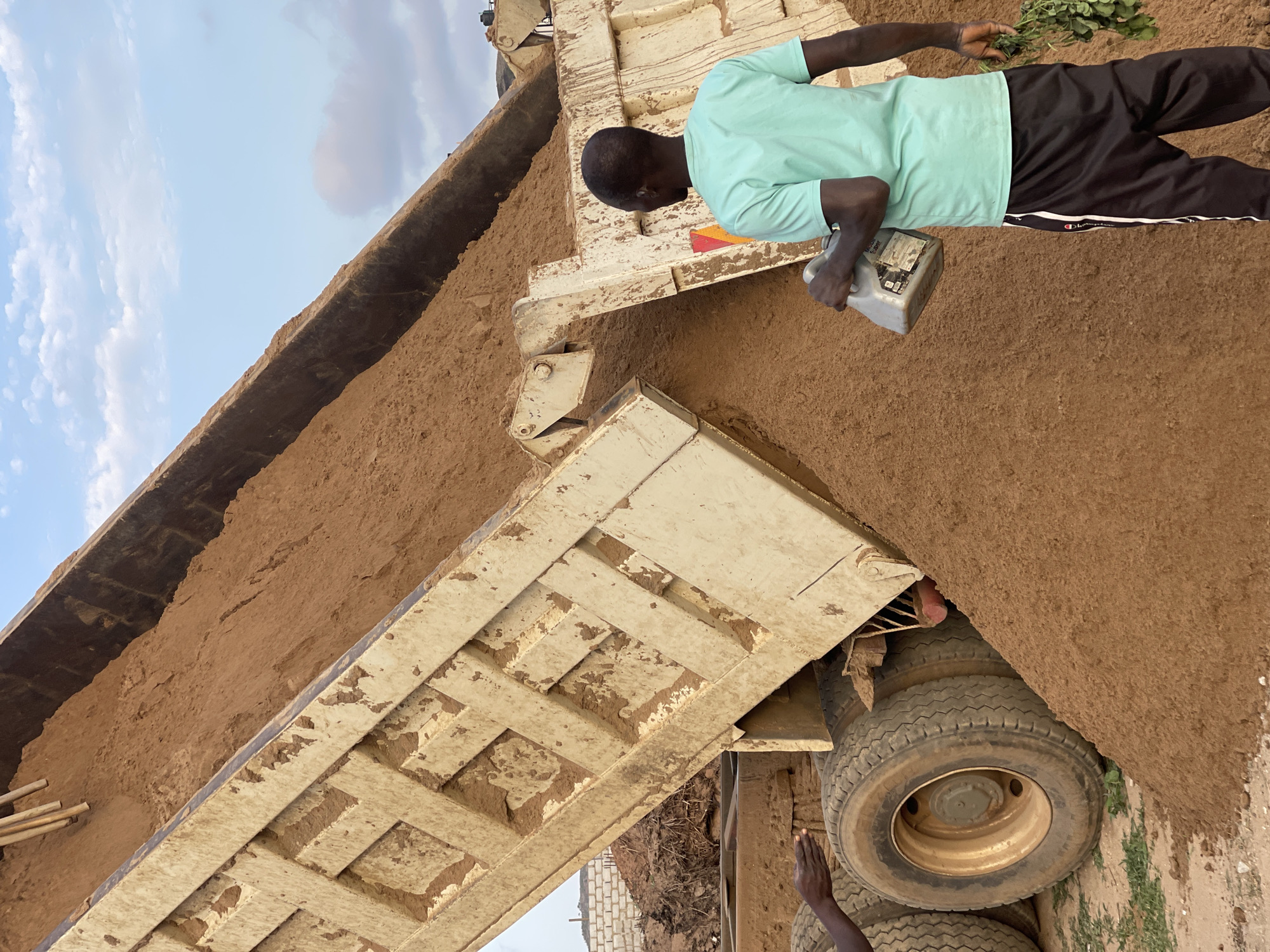 In Angwan Sayawa, Nigeria, a dump truck is tilted to unload dirt. A Black man wearing a mint-green t-shirt and dark pants with a white stripe, is pouring liquid from a light grey container onto the dirt load and holding green foliage in his other hand. The tires of the truck and a brick wall are visible. The sky is visible in the background.
