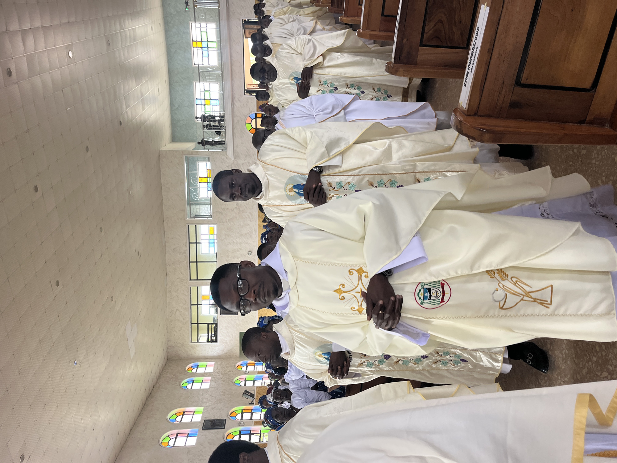 A group of men in white and cream robes stand in a church, likely during a religious ceremony in Vom, Nigeria. The men are mostly of African descent, with some wearing glasses. They appear to be priests or clergy members, as indicated by their attire. The church interior features high ceilings, stained-glass windows with colorful panes, and rows of wooden pews. The lighting suggests it is daytime, with natural light entering through the windows and some artificial lighting from above. The overall atmosphere is solemn and formal. The men are standing in a procession or formation, facing forward. Their hands are clasped in front of them. The focus is on the individuals in the foreground, who are well-dressed in ceremonial robes. The background shows more people and the architectural elements of the church. A sign on the pew in the foreground reads "CMO/ZUMUNCI MAZA RESERVED FOR".