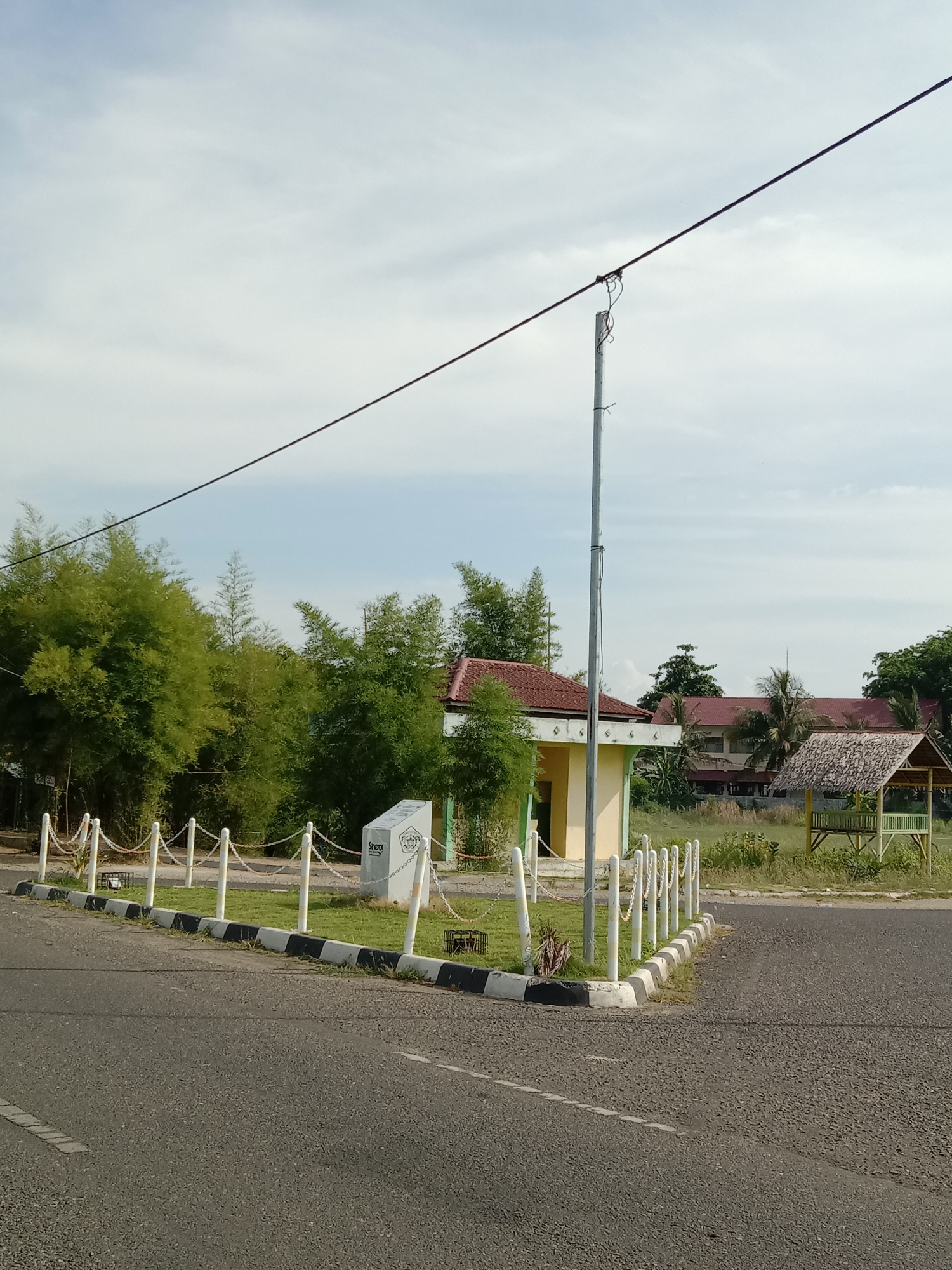 The image depicts a roadside scene in Lhokseumawe, Indonesia, with a road in the foreground splitting in two directions. Along the edge of the road is a small, landscaped area enclosed by white posts connected with chains. A monument or sign stands within this area.
Behind this area are several buildings, including a small, yellow building with a red tile roof, and a gazebo-like structure with a thatched roof. Various trees and vegetation are also present. An overhead power line runs diagonally across the upper part of the frame, supported by a metal pole. The sky is overcast with light clouds.