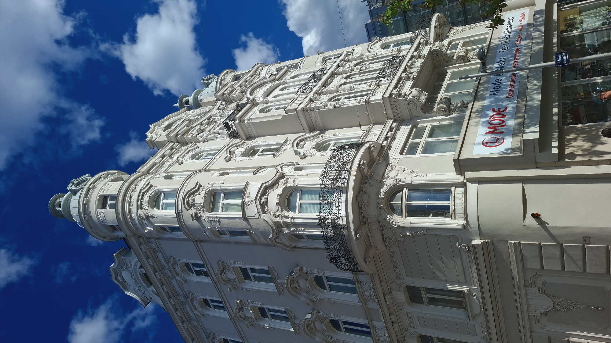 A vertical shot shows the facade of a light-colored, ornamented building against a background of a bright blue sky with white clouds. The building features elaborate architectural details, including arched windows, decorative moldings, and iron balconies. A corner tower with a dome sits at the left. A vertical sign on the right side displays the word "MODE" in red and white and further text. The building is the Mode & Mehr on Mariahilfer Straße in Vienna, Austria.