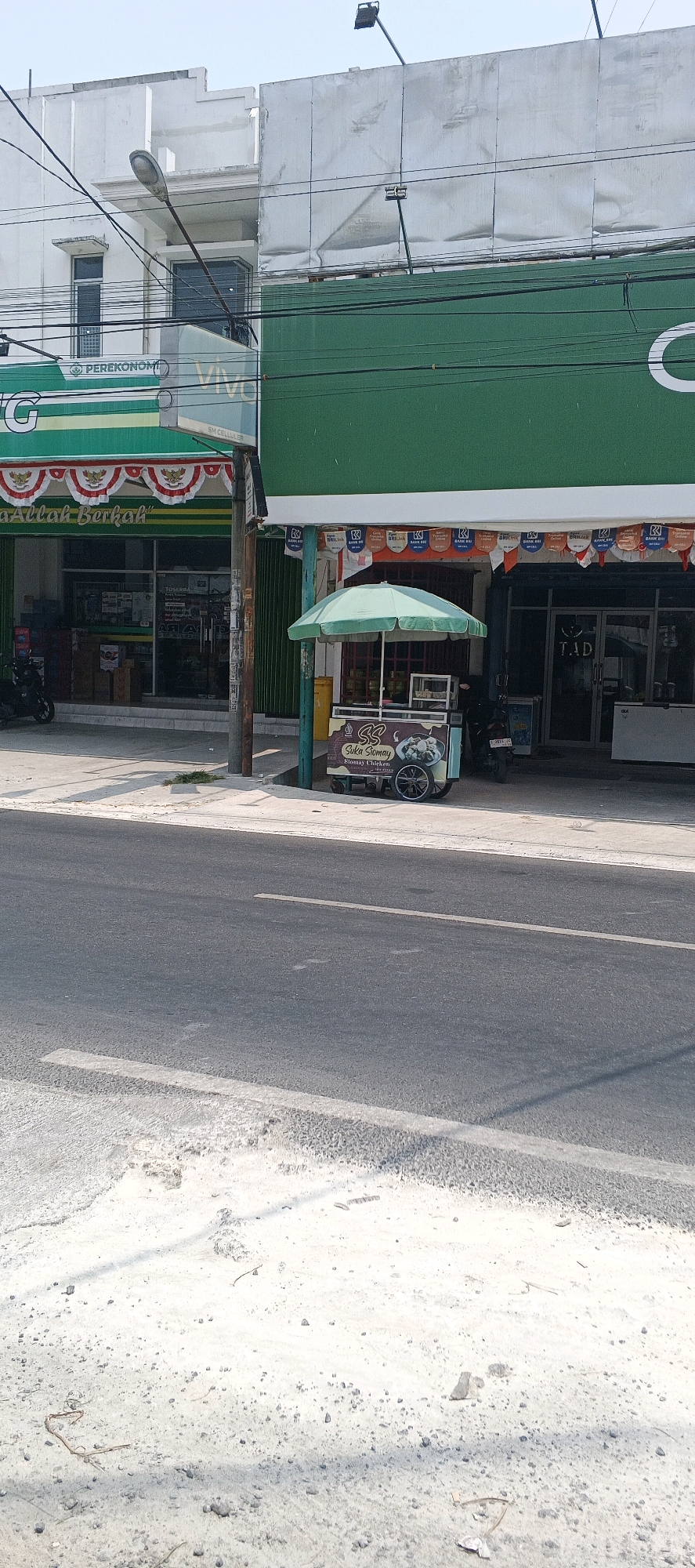 The image depicts a street scene in Karanggeneng, Indonesia. In the foreground, a gray asphalt road with white lane markings is visible, covered in some scattered gravel and debris. The middle ground features a sidewalk adjacent to a row of shops. To the left, a green awning with white text that includes "PEREKONOM" is seen, above a storefront. Next to it, a green sign with white text, possibly advertising a phone service, is present. Further to the right, a food cart with a green umbrella is parked. The cart has a sign that reads "Suka Siomay" and "Siomay Chicken." Behind the cart, a dark storefront with the letters "T.A.D." is visible, with a closed roll-up door. Electrical wires are strung across the scene, connecting to poles. In the background, a multi-story building with a white facade and large, reflective surfaces is present, topped by a blank billboard. The sky is overcast. No people are clearly visible in the image.