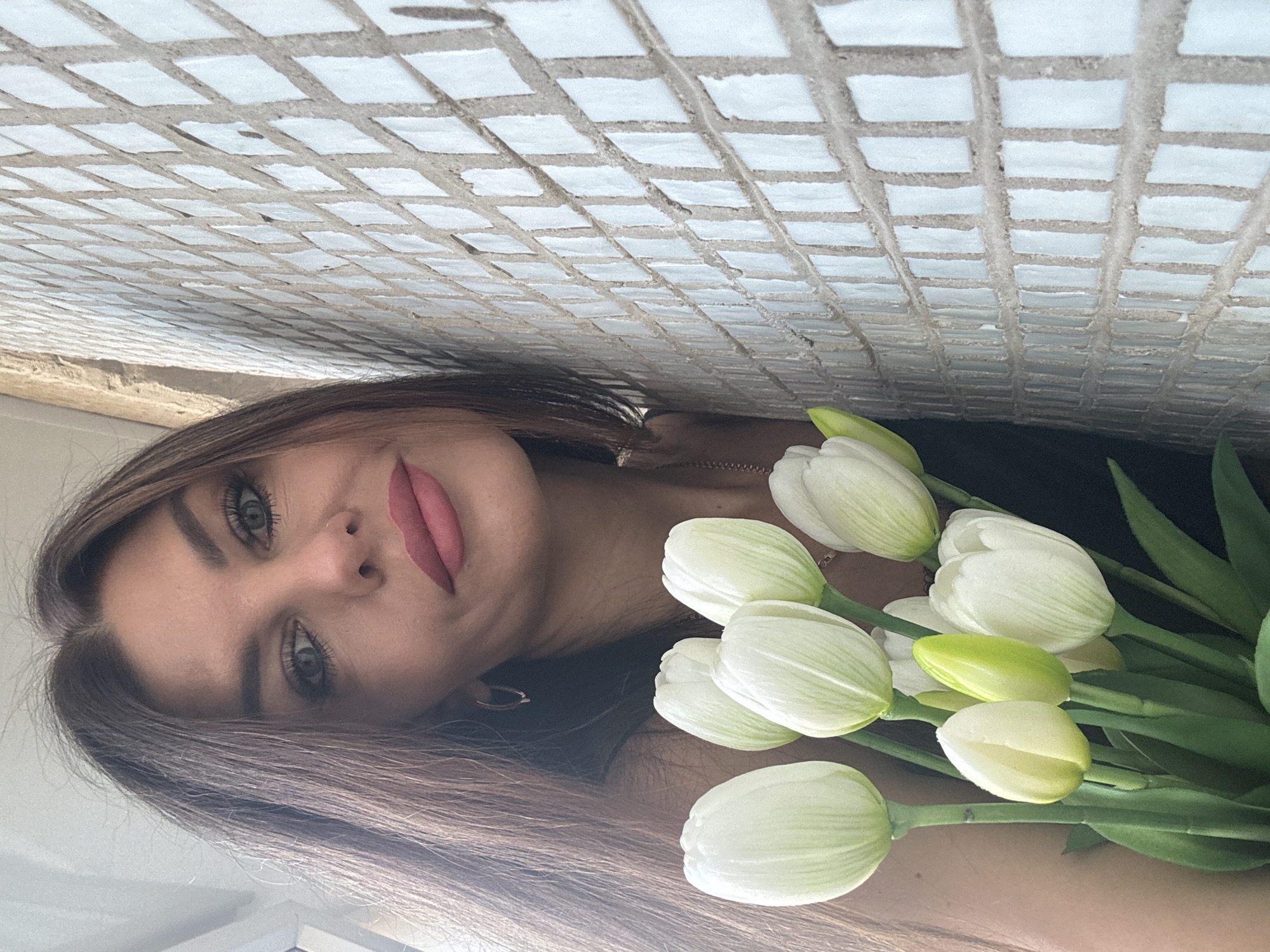 A woman with long brown hair, pink lipstick, and light eyes is holding a bouquet of white tulips. The background appears to be a tiled wall. The location is Chaikovsky, Russia.