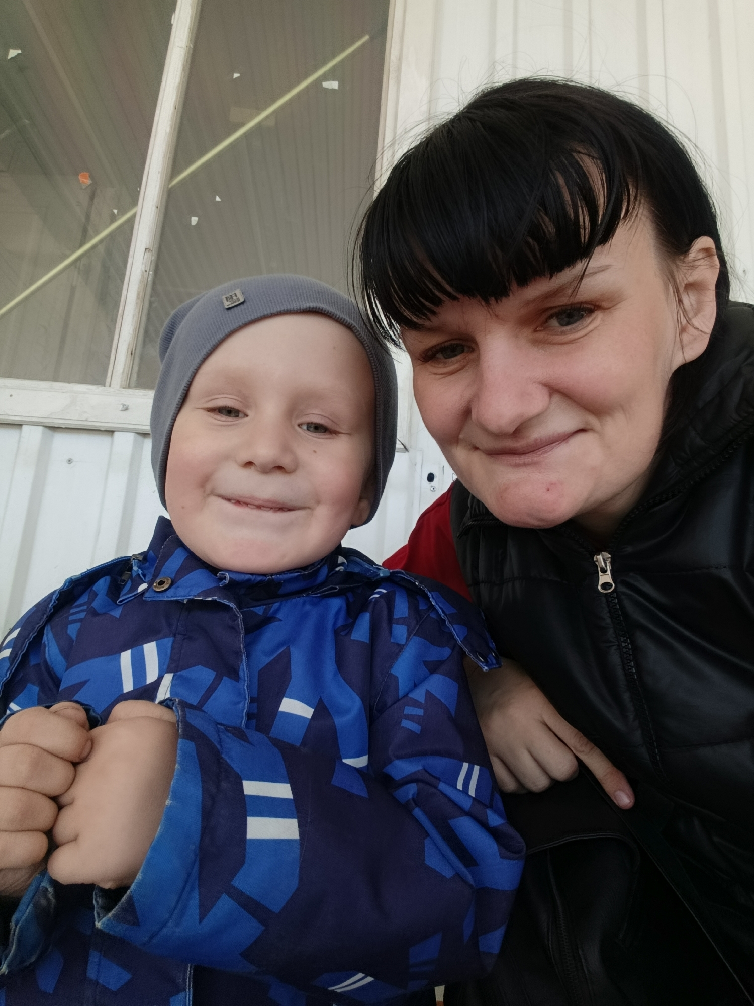 A medium shot photograph depicts a woman and a young boy posing for a selfie. The boy, on the left, wears a grey knit cap and a blue winter coat with white geometric patterns. He is smiling broadly, with his hands clenched into fists inside his sleeves. The woman, on the right, has dark hair with bangs and wears a black leather jacket over a red garment. She is also smiling slightly. Behind them is a white, corrugated metal wall with a window frame above. The location is likely in the city of Chaikovsky, Russia. The image conveys a sense of warmth and closeness between the two individuals.