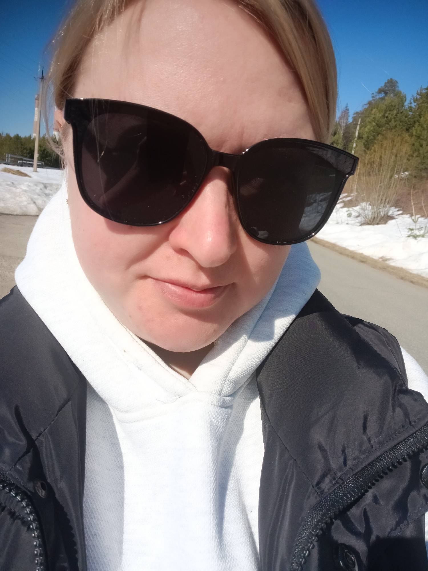 A selfie of a blonde woman wearing large black sunglasses, a white hoodie, and a black vest. The background is a partially snowy landscape. A road is visible to the right, and trees and a blue sky are visible in the distance. A utility pole is visible on the left. The location is Chaikovsky, Russia.