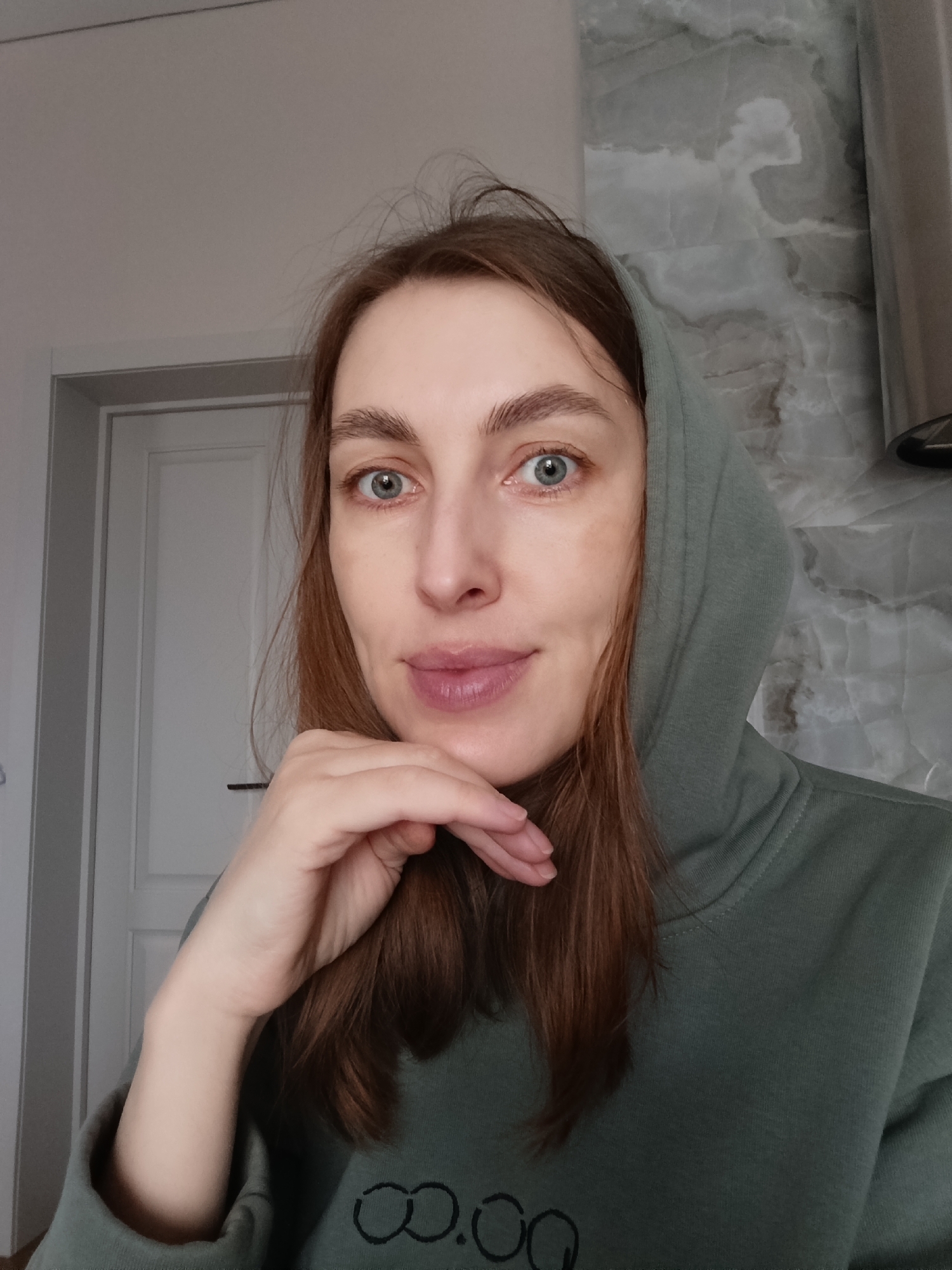 A woman with fair skin, light brown hair, and blue-green eyes poses for a selfie. She is wearing an olive green hooded sweatshirt with a logo on the lower front. Her left hand is raised, resting her chin on her fingertips. The background includes a white door on the left, and a gray marbled wall on the right.
There is no information to suggest that this image was taken in Chaikovsky, Russia.