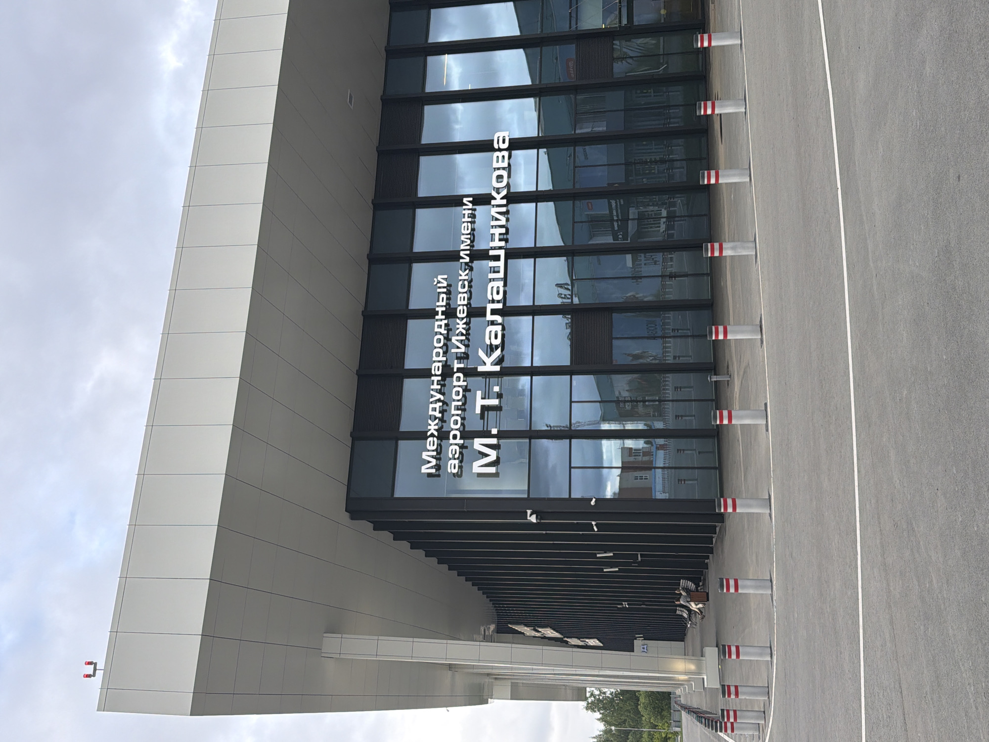 This image depicts the Izhevsk International Airport terminal, located near Staroe Mart'yanovo, Russia, under an overcast sky. The modern building features a prominent, expansive white cantilevered roof and a facade primarily composed of large, dark-tinted glass panels. On the far left corner of the roofline, two red obstruction lights are visible. Large white Cyrillic text on the glass facade reads "Международный аэропорт Ижевск имени М. Т. Калашникова," which translates to "Izhevsk International Airport named after M. T. Kalashnikov." The building's left section incorporates vertical dark slats or panels between the glass segments.
In the foreground, a dark asphalt surface with a single white painted line extends across the bottom of the frame. A curved line of approximately ten light-grey cylindrical bollards, each topped with red and black stripes, separates the roadway from the building's pedestrian area. Several smaller, solid grey posts are also interspersed among the striped bollards. Two individuals are seated on a dark bench to the left of the main entrance, beyond the bollards. One person is dressed in darker attire, and the other in lighter clothing; a third person is partially visible further down the bench. Green trees and foliage are visible in the far background on the left, suggesting the surrounding natural environment.