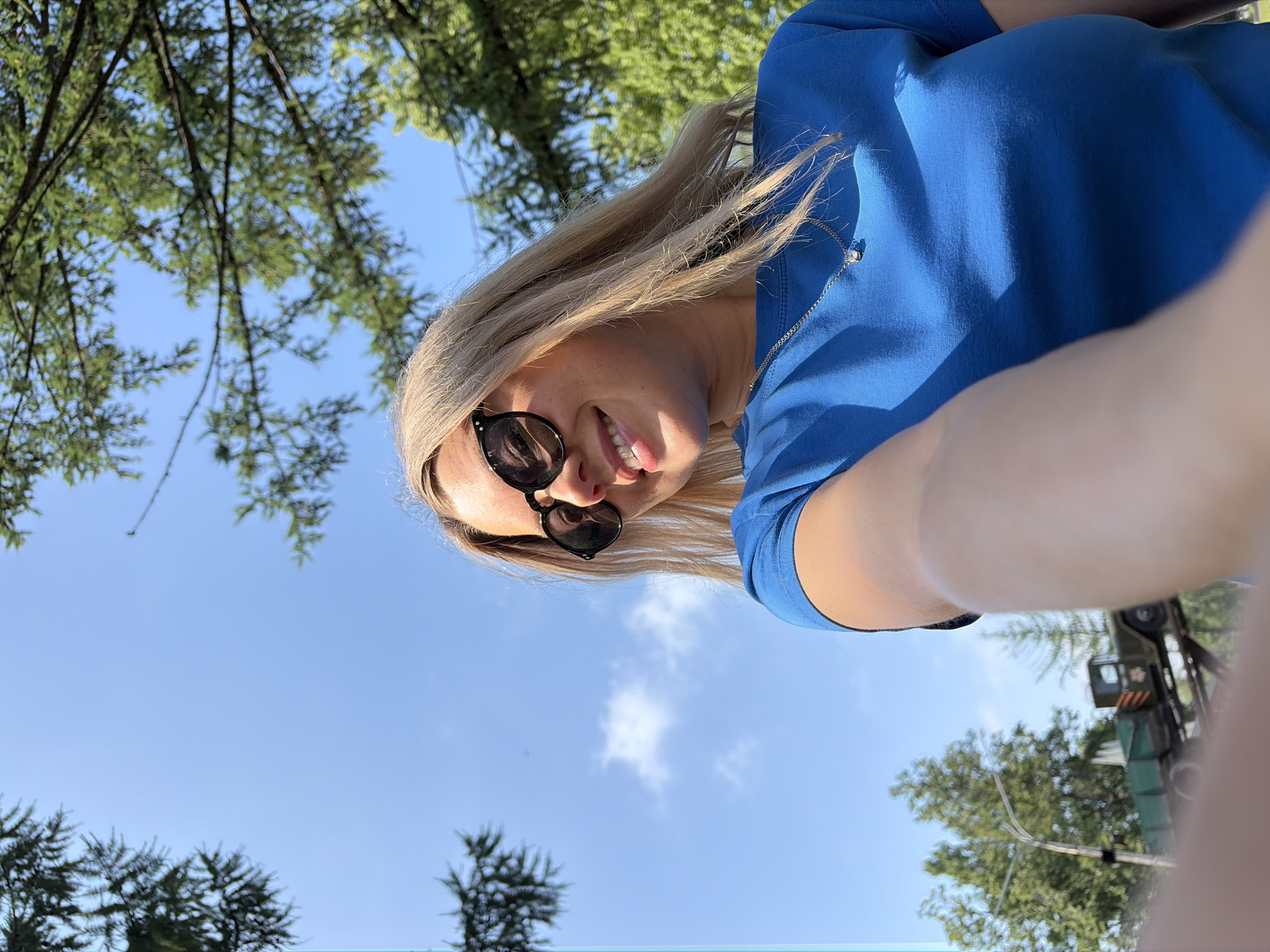 A young woman with blonde hair and sunglasses smiles as she takes a selfie outdoors. She is wearing a blue t-shirt and a silver necklace with a small pendant. In the background, there are trees and a clear blue sky with a few clouds. To the left of the woman, in the distance, a green truck is visible. The image appears to have been taken on a sunny day. The location is the city of Chaikovsky, Russia.