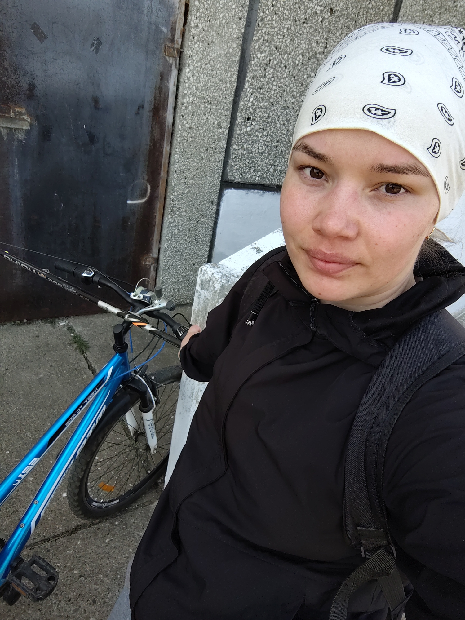 A person wearing a white bandana and a black jacket is standing next to a blue bicycle on a concrete surface, likely a sidewalk or a road, with a gray concrete building in the background. A dark-colored, possibly metal, door or structure is also visible behind the bicycle. The person is holding the handlebars of the bicycle. The location is identified as Марковский, Russia.