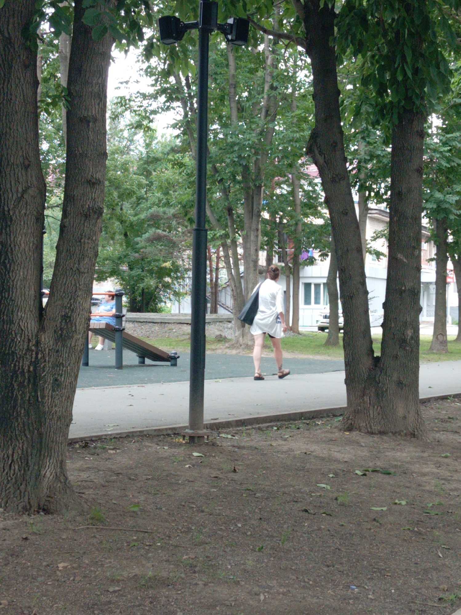 A woman in a white tunic and black shorts walks on a paved path in a park. She is carrying a black tote bag over her left shoulder. A lamppost with two black fixtures is to her right. In the background, a person in a blue shirt is sitting on a park bench. Trees and buildings are visible in the distance. The ground is covered in dirt and leaves.