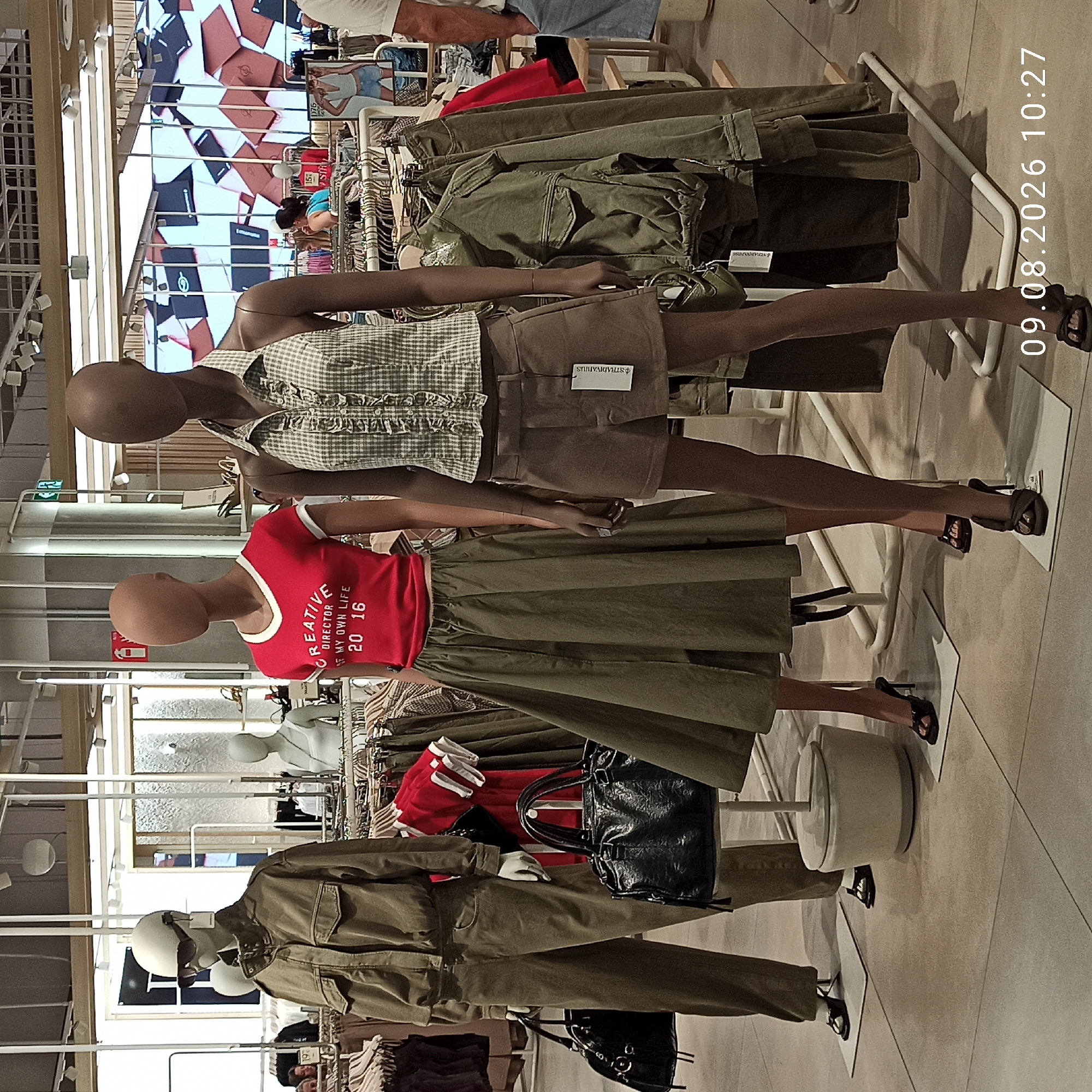 The image shows a retail display within a clothing store in Molfetta, Italy. Two mannequins are positioned prominently in the foreground. The mannequin on the left is wearing a khaki jumpsuit and sunglasses, holding a black handbag. The mannequin on the right is dressed in a checkered halter top and brown shorts, also holding a handbag. Behind them, clothing racks are filled with various garments, including jackets and shirts. In the background, a large digital display shows a collage of magazine covers and product advertisements. Several shoppers are visible in the middle ground, browsing the merchandise. The store appears to be brightly lit with a modern, clean aesthetic. The timestamp "09.08.2026 10:27" is visible in the bottom right corner of the image.