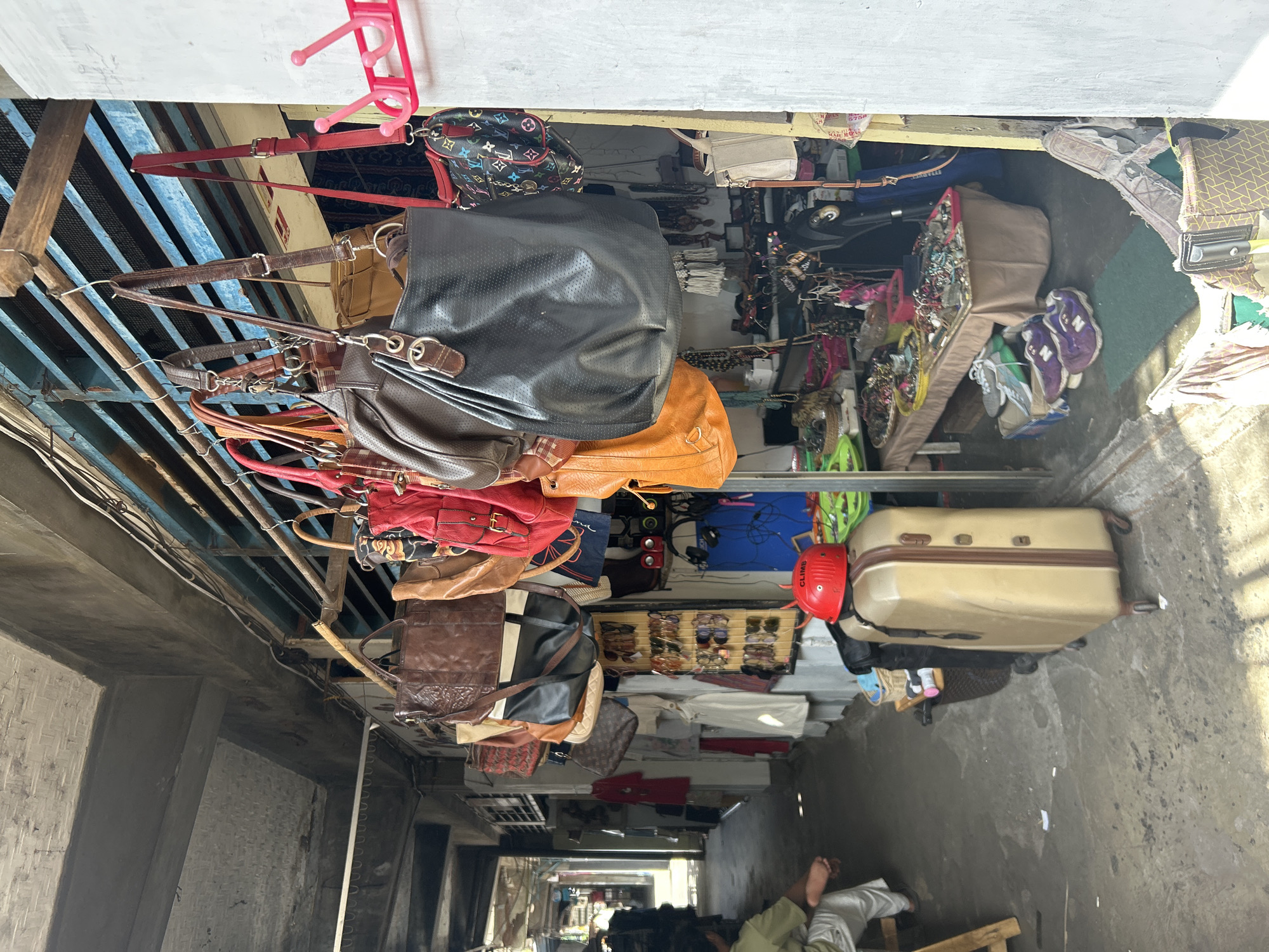 The image depicts an outdoor market stall in Denpasar, Indonesia. The stall is filled with various bags and accessories. In the foreground, a tan suitcase with brown trim sits on the ground, topped by a red helmet. A person in a light-colored shirt and pants is partially visible on the left, seated on a wooden bench. The stall itself features an array of handbags hanging from a metal rack, including brown leather bags, red bags, a black textured bag, and a bag with a multi-colored monogram pattern. Below the hanging bags, shelves are stocked with jewelry, sunglasses, and other small items. The background reveals a narrow pathway lined with more stalls, suggesting a bustling market environment. The structures in the background appear to be made of concrete and metal.