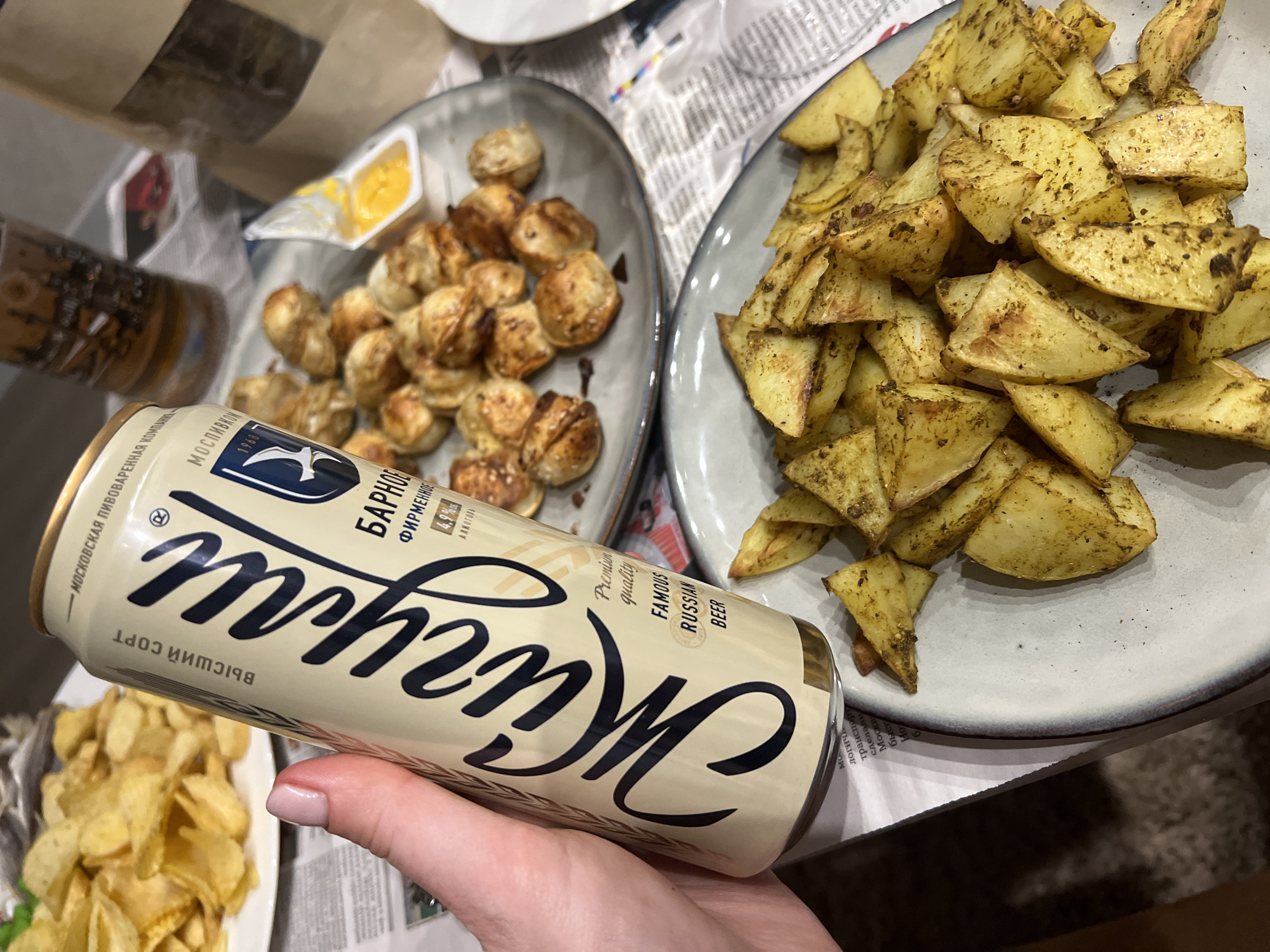 An overhead, close-up shot captures a casual meal featuring beer and snacks. A hand is holding a light-colored can of "Жигули" beer, prominent in the foreground. The can is adorned with Cyrillic and English text, including "Premium quality," "FAMOUS RUSSIAN BEER," and "4,9% АЛКОГОЛЬ" (4.9% ALCOHOL). Other legible text on the can includes "БАРНОЕ" (BARNOE), "ФИРМЕННОЕ" (FIRMENNOE), "ВЫСШИЙ СОРТ" (HIGHEST GRADE), "МОСКОВСКАЯ ПИВОВАРЕННАЯ КОМПАНИЯ" (Moscow Brewing Company), and the year "1968".
On the table, two plates of food are visible. To the left of the beer can, a plate holds numerous small, golden-brown fried dumplings or pelmeni, accompanied by a small plastic container of yellow sauce, likely cheese or mustard. To the right, a plate is piled high with seasoned, roasted or fried potato wedges, showing a golden-brown crust and a dusting of green herbs or spices.
The table is covered with newspapers, serving as a makeshift tablecloth, with snippets of Russian text visible, such as "логича", "транспор", "сделали", and "Москв". The lighting suggests an indoor setting, possibly evening, emphasizing the warm tones of the food and beer can. The scene evokes a relaxed atmosphere, typical of a snack or meal enjoyed at home.