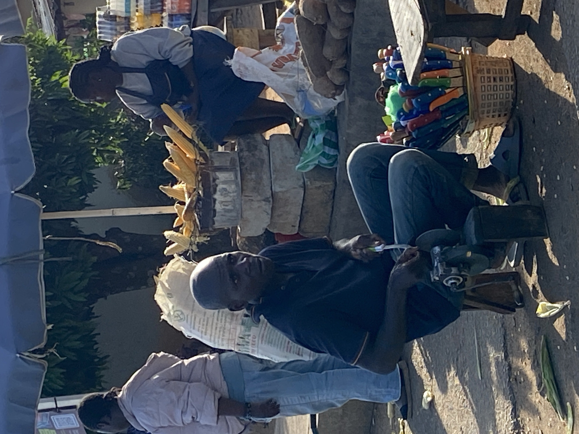 The image captures a street scene in Bukuru, Nigeria. A man in a dark blue shirt and jeans is seated, sharpening knives using a grinding machine placed on a low stool. A basket filled with colorful knives sits beside him. Behind him, another person, presumably a vendor, is attending to corn on the cob displayed on a makeshift stall constructed from concrete blocks. A bag of yams is visible, leaning against the stall. Another person is standing nearby, wearing a light pink shirt and blue jeans. The environment suggests an outdoor market setting.