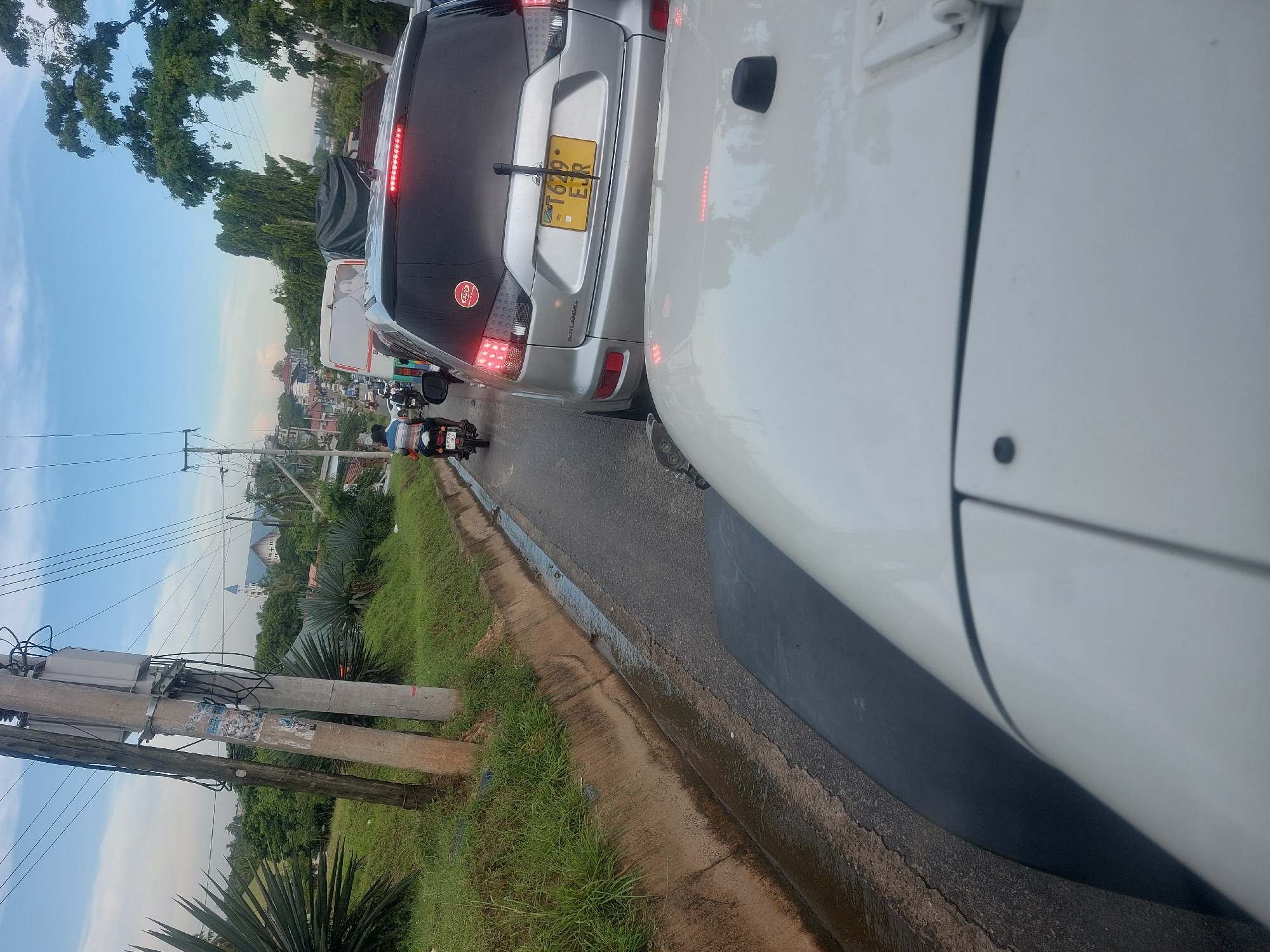The image shows a traffic jam on a road in Dar es-Salaam, Tanzania. The view is from inside a white vehicle, looking at the rear of a silver SUV with the Tanzanian license plate T629 EUR. Other vehicles, including a truck and motorcycles, are visible in the distance. The sky is blue with some clouds. Utility poles and green vegetation line the side of the road. A church steeple is visible in the background.