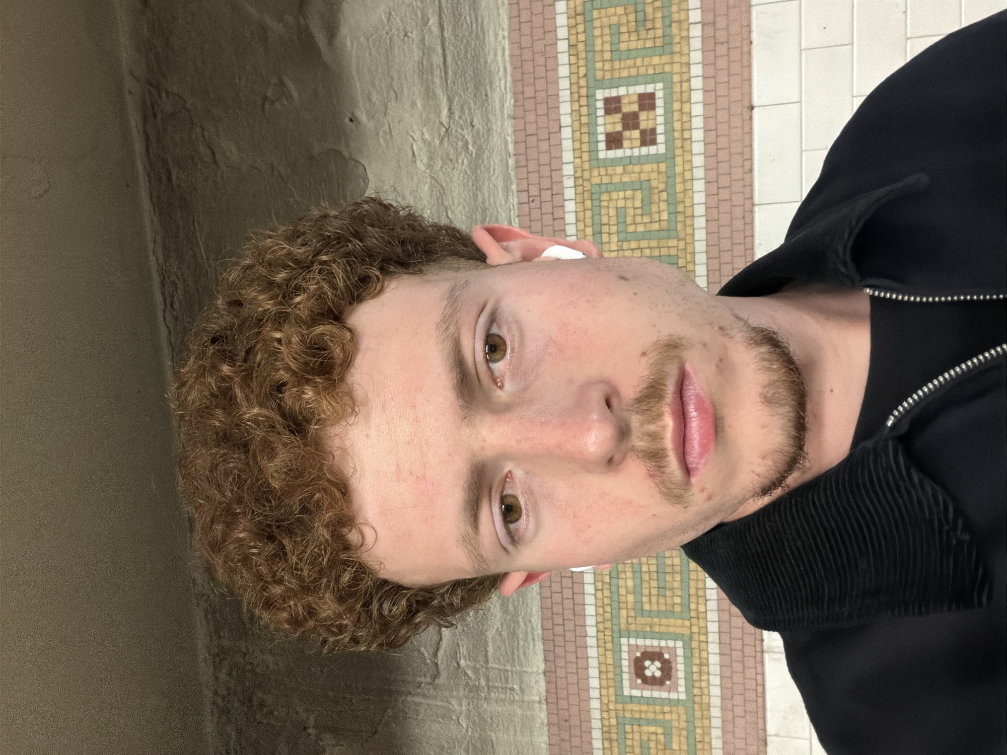 A close-up, eye-level shot features a Caucasian man with curly reddish-brown hair, light skin, and hazel eyes. He has a light mustache and beard. He is wearing a black jacket with a silver zipper. An earbud is visible in his left ear. The background includes a textured beige wall on the left and a decorative tiled surface with a geometric pattern on the right. The tiled pattern consists of small square tiles in alternating colors of brown, white, beige, and green. The tiles suggest that the location may be the New York City Subway system, in New York, United States of America.