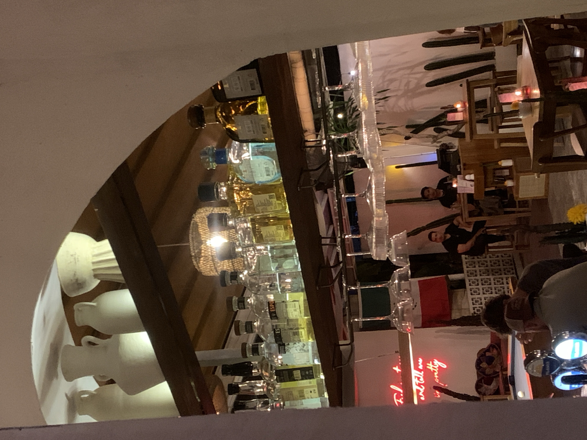 The image depicts the interior of a dimly lit bar or restaurant in North Kuta, Indonesia. The scene is framed by an arched opening, with a wooden shelf above holding several large, cream-colored decorative vases or amphorae. Below this shelf, another wooden shelf displays an assortment of liquor bottles, primarily tequila, with various labels and colored liquids. Hanging glassware is visible below this shelf.
In the mid-ground, a pool table with a white ball is partially visible. To the left of the pool table, neon pink text on a white background reads "Feel the..." and then "and tell me you're pretty". There are two men seated at a table to the right, and another man is seated on a stool in the background. Further to the right, several small tables with lit candles are visible, set against a backdrop of minimalist wall art featuring stylized cacti. The overall atmosphere suggests a relaxed, possibly nighttime, social setting.