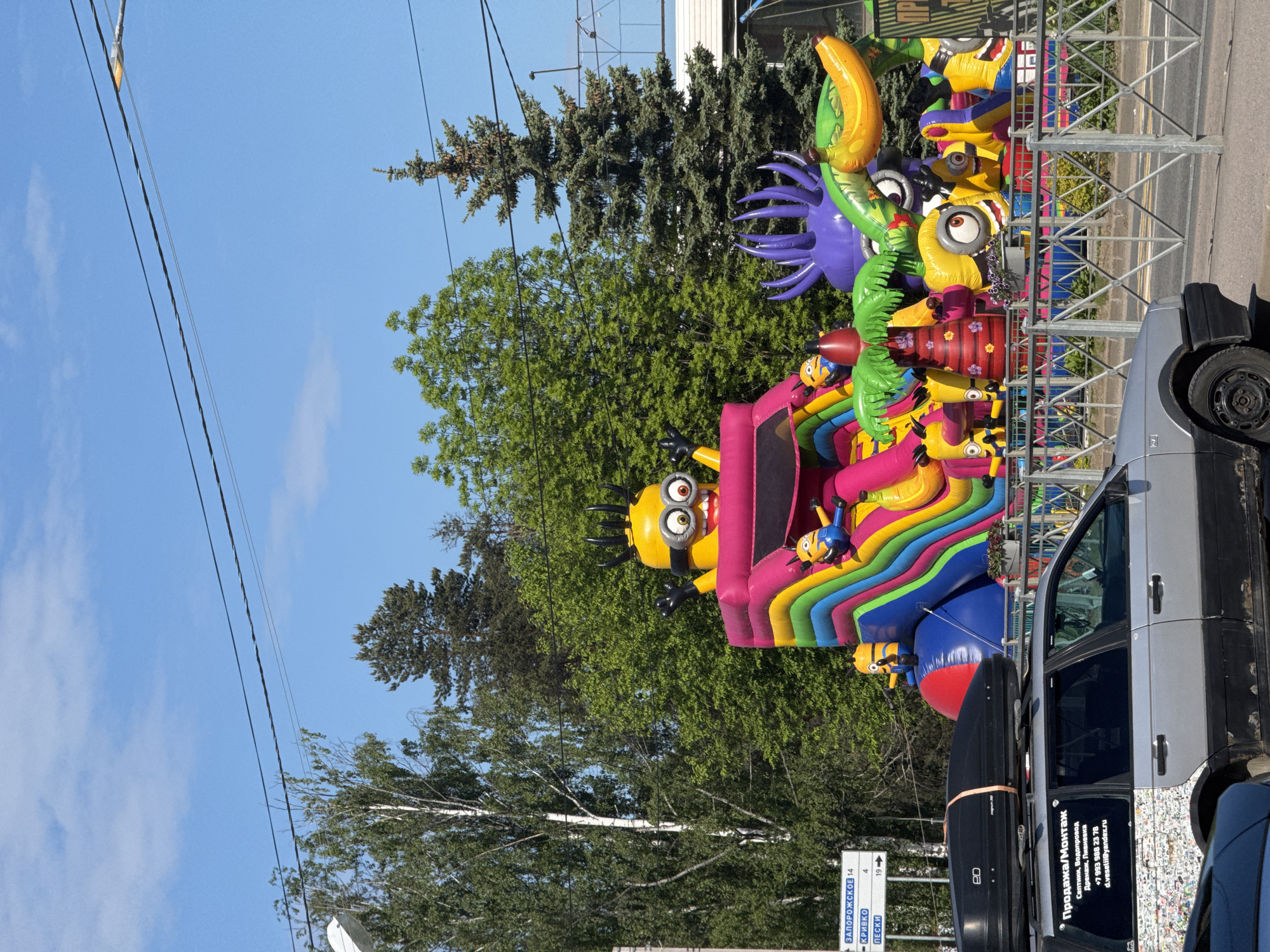 An outdoor scene in Sosnovo, Russia, features a vibrant inflatable bouncy castle complex designed with numerous Minion characters, suggesting a children's event or entertainment setup. The main inflatable structure is multi-colored, with yellow and pink elements, and several Minion figures are integrated into its design, some appearing to slide or climb. Other large inflatables include a purple octopus-like creature, a yellow banana, and additional Minions.
To the right, a silver/grey car, possibly a van, is parked. Its side is adorned with text in Cyrillic, which reads "Продажа/Монтаж Септики, Водопровод Дренаж, Ливневка" (Sales/Installation of Septic tanks, Water supply, Drainage, Storm drains), followed by a phone number "+7 993 988 23 78" and an email "d.veselii@yandex.ru". A black roof box is mounted on top of the car, which also bears an "eD" logo. A metal fence separates the inflatables from the background.
The background consists of lush green trees, including both tall coniferous trees and leafy deciduous trees, under a clear blue sky with a few faint clouds. Power lines stretch across the upper part of the image. In the lower left, a blue and white road sign is visible, displaying Cyrillic place names with distances: "ЗАПОРОЖСКОЕ 14", "КРИВКО 4", and "ПЕСКИ 19". The overall impression is a sunny daytime scene with colorful, playful elements.