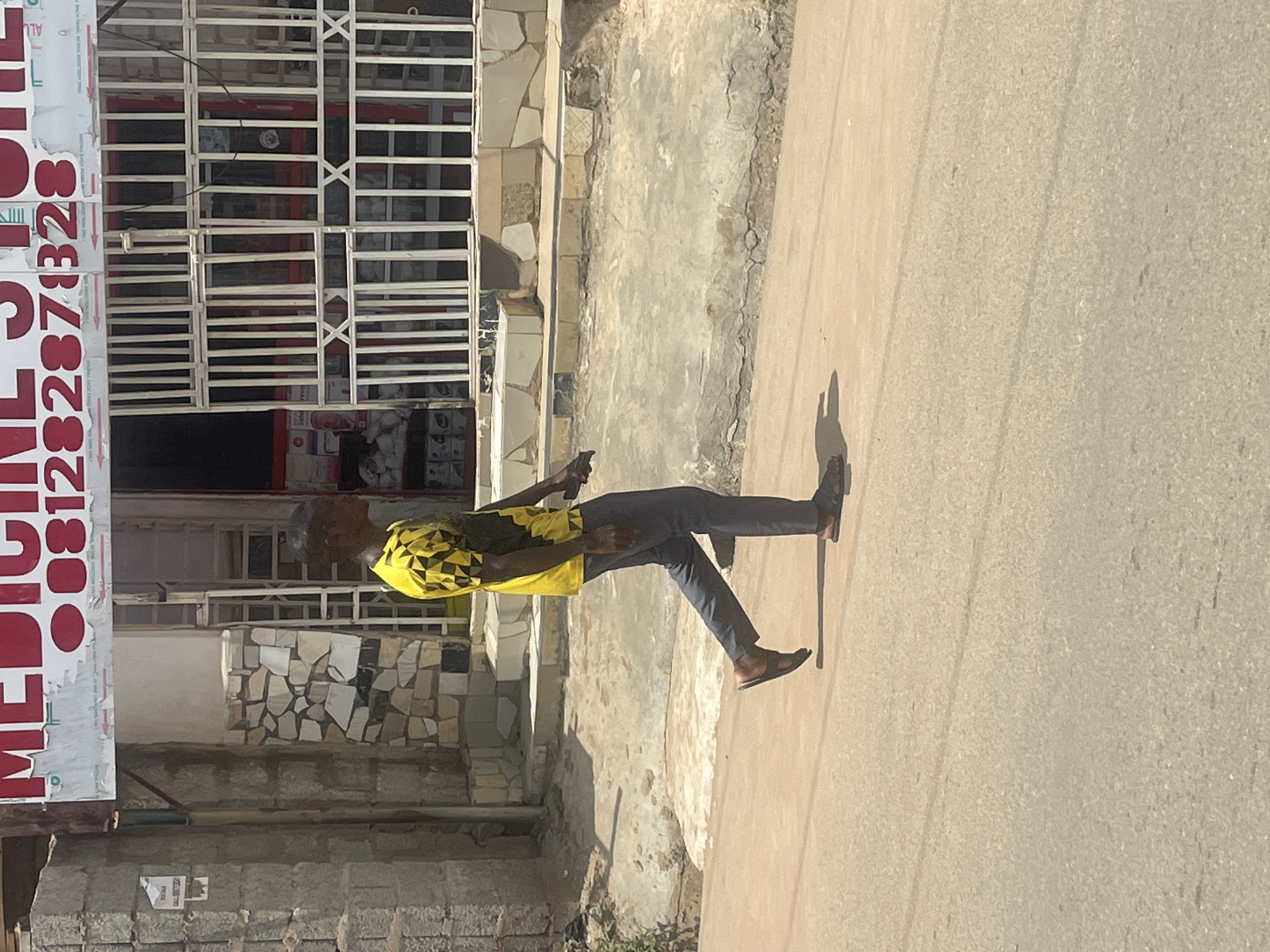A man in a yellow and black patterned t-shirt and gray pants is walking on a paved street in front of a medicine store. The medicine store has a metal gate and shelves visible inside. A sign above the store reads "MEDICINE STORE" with a phone number. The street is dusty and cracked, with some debris on the side. The environment appears to be an urban setting in Abuja, Nigeria, given the style of the building and the clothing of the person.