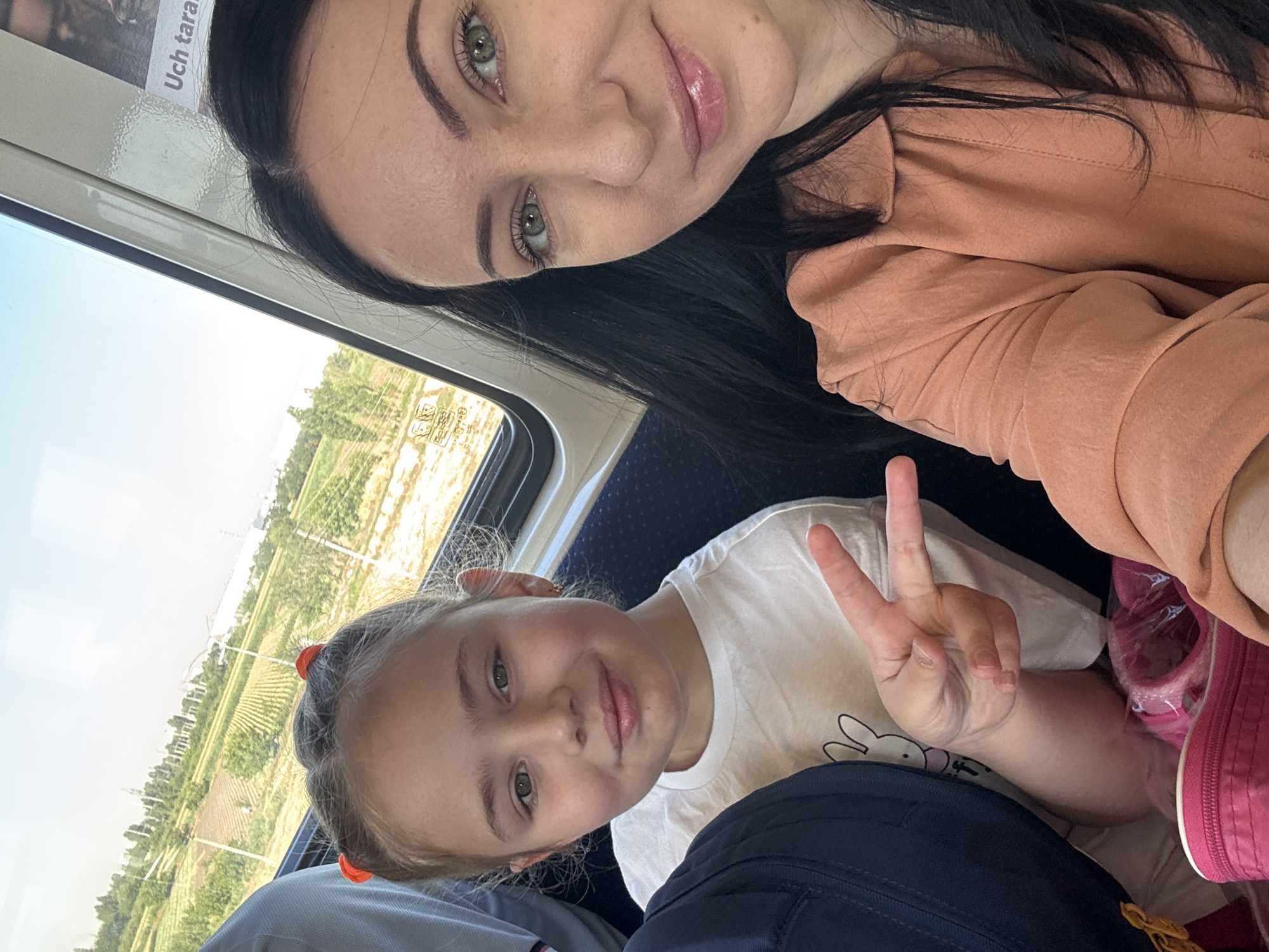 This media file features two people, a woman and a young girl, likely mother and daughter, seated on a train. The woman is in the foreground, her face partially visible as she takes a selfie. She has dark hair and green eyes, and is wearing an orange shirt. The young girl is next to her, also looking towards the camera. She has blonde hair with orange hair ties and is wearing a white t-shirt with a bunny graphic. She is holding up two fingers in a peace sign. The background shows a view of a rural landscape with fields and trees, suggesting the train is traveling through the countryside. The lighting indicates it is daytime, likely sunny. The image is a snapshot of a moment during a train journey. No discernible text is visible within the frame itself, though a cropped section shows what might be part of a newspaper or advertisement with text that is too blurry to read. The overall mood is casual and cheerful.