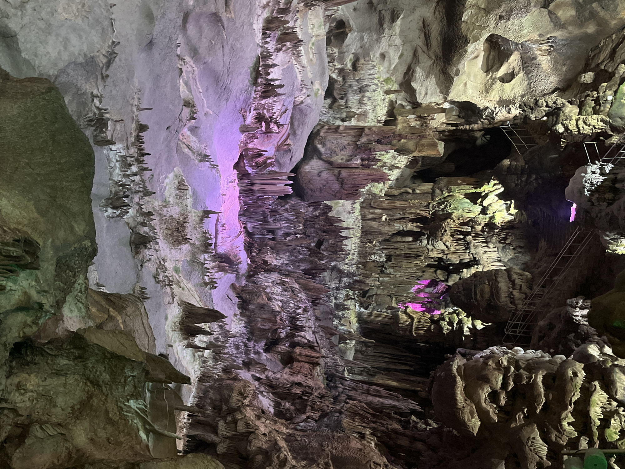 The image depicts the interior of a cave, characterized by stalactites and stalagmites. The cave is illuminated by artificial lights, with sections bathed in purple and green hues, creating a dramatic and somewhat surreal atmosphere. Narrow metal staircases are visible, suggesting a pathway or viewing area for visitors within the cave. The rock formations are varied in texture and shape, with some appearing smooth and others rough and jagged. The overall scene conveys a sense of natural wonder and the impressive scale of geological formations. There is no visible text in the image. The time of day and weather are not discernible as the scene is entirely underground. No people are visible, nor is there any indication of an event taking place beyond exploration or tourism.