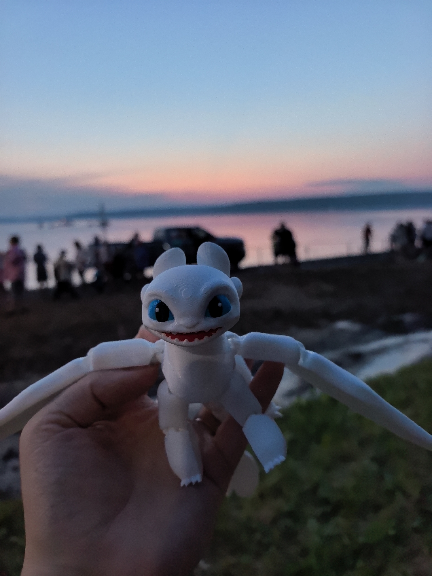 A white, humanoid dragon figurine with blue eyes and red accents is held in a human hand, positioned in the foreground. The figurine's wings are outstretched to the sides, and its mouth is open, revealing red gums and white teeth. The figurine is being held against a blurred background of a body of water under a twilight sky painted with hues of pink, orange, and blue. Along the shoreline, silhouetted figures of people and several vehicles are visible. The environment appears to be a natural setting, possibly a beach or a park, near a body of water. The image was taken at dusk or dawn. There is no specific information to confirm the location as Osa, Russia.