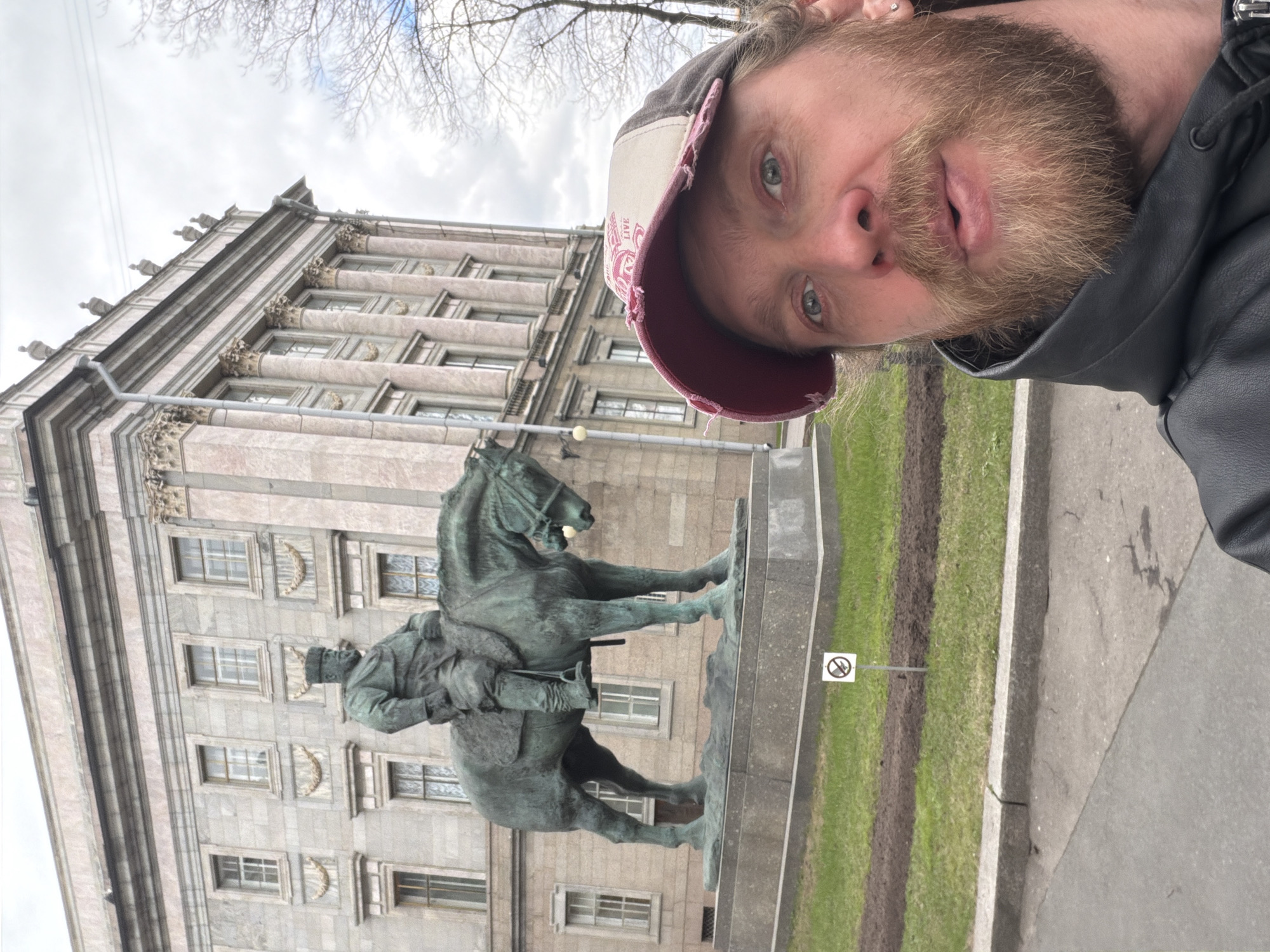 The image is a vertically oriented selfie taken by a man, capturing himself, an equestrian statue, and a grand historical building in Saint Petersburg, Russia.
In the foreground, a man with a light brown beard and mustache and light-colored eyes looks directly at the camera. He is wearing a baseball cap with a dark brim and a lighter crown, and a dark jacket. His expression is neutral.
Behind the man, a prominent bronze equestrian statue depicts a historical figure, possibly a military leader or emperor, on horseback. The horse stands with one leg slightly raised. Further in the background is a large, multi-story building featuring classical architectural elements such as numerous rectangular windows, pilasters, cornices, and detailed decorations, suggesting it is the Mariinsky Palace. Bare tree branches are visible against an overcast, grey sky.
The scene is an urban outdoor setting. To the right of the statue, a grassy area with some muddy patches is visible, separated from a paved surface by a curb. A white sign with a red circle and a diagonal line through it, indicating a prohibition (e.g., "no entry" or "no walking"), is planted in the grass. The overall impression is of a visitor documenting their presence at a significant historical landmark in Saint Petersburg on an overcast day.