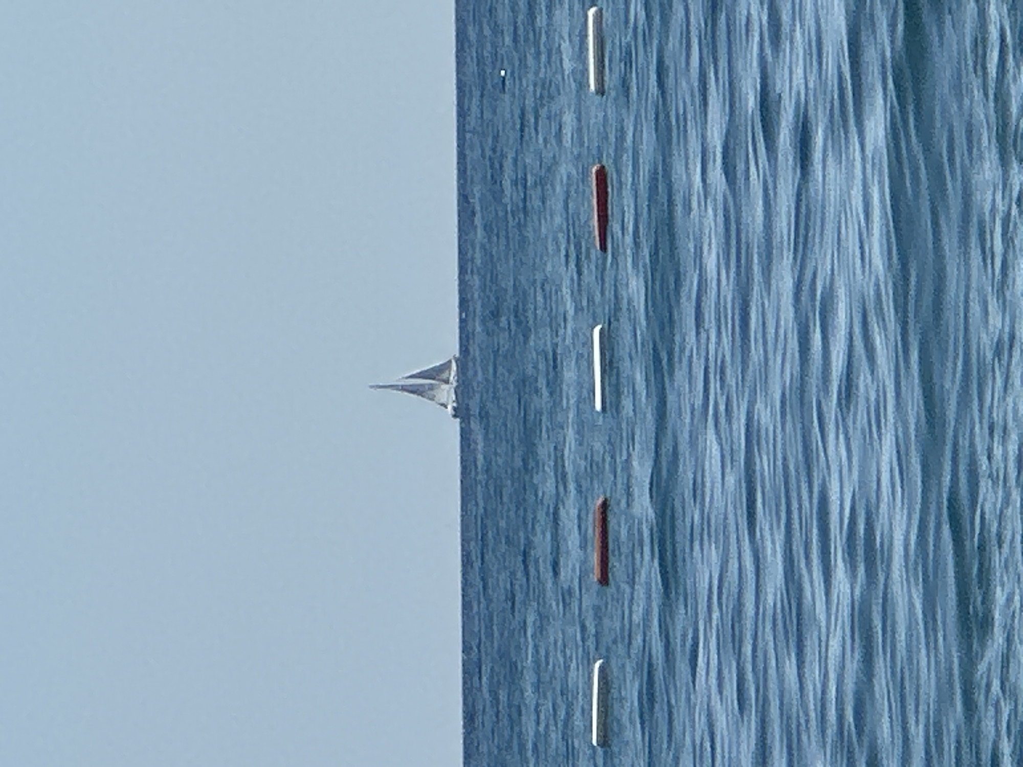 The image depicts a serene seascape with a clear, pale blue sky meeting a deep blue ocean. In the distance, a single sailboat with two sails is visible against the horizon. Closer to the foreground, a line of buoys floats on the water, marking a boundary. There are three visible buoys: two white rectangular buoys are spaced apart, with a single dark red rectangular buoy positioned between them. The water is calm, with gentle ripples reflecting the light. The overall impression is one of peacefulness and open space. The location is specified as Boreti, Montenegro.