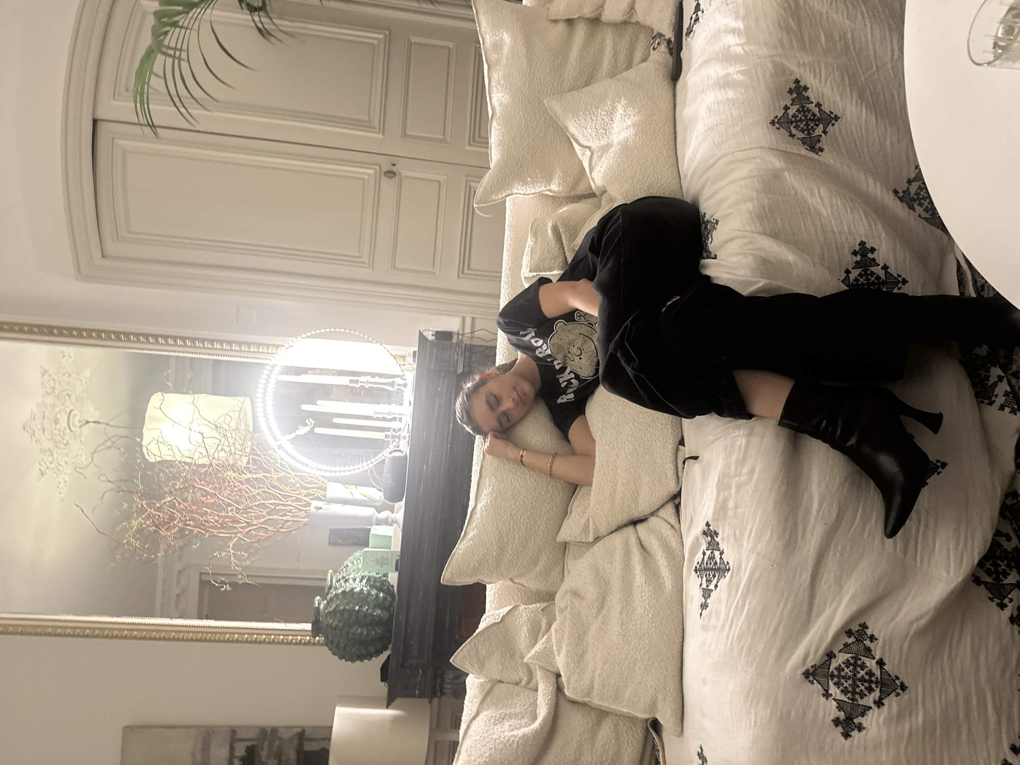 A young woman rests on a sofa in an interior setting. She is lying on her side, face visible, wearing a black graphic tee, black pants, and black heeled boots. The sofa is covered with light-colored pillows and a patterned duvet. A large mirror reflects a lamp and interior décor, including a fireplace. A potted plant is also visible. A white, arched doorway can be seen in the background. The scene takes place in Paris, France.