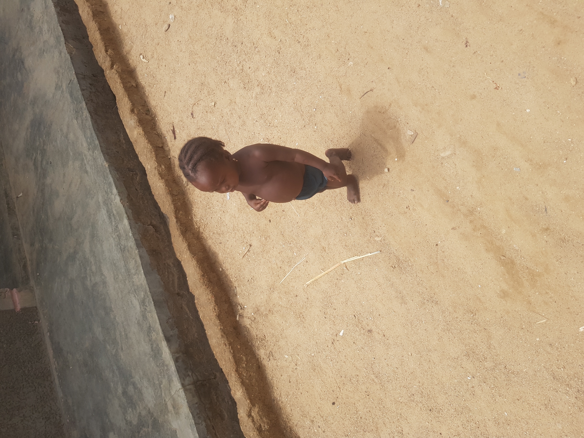 A young child stands barefoot on a sandy surface next to a concrete structure. The child has dark skin, braided hair, and is wearing dark-colored shorts. The child is facing towards the concrete structure. The sand is light brown and covers the ground. A few small pieces of debris are scattered on the sand. The concrete structure is gray and appears to be a wall or curb. The location appears to be outdoors in Dutsinma B, Nigeria.