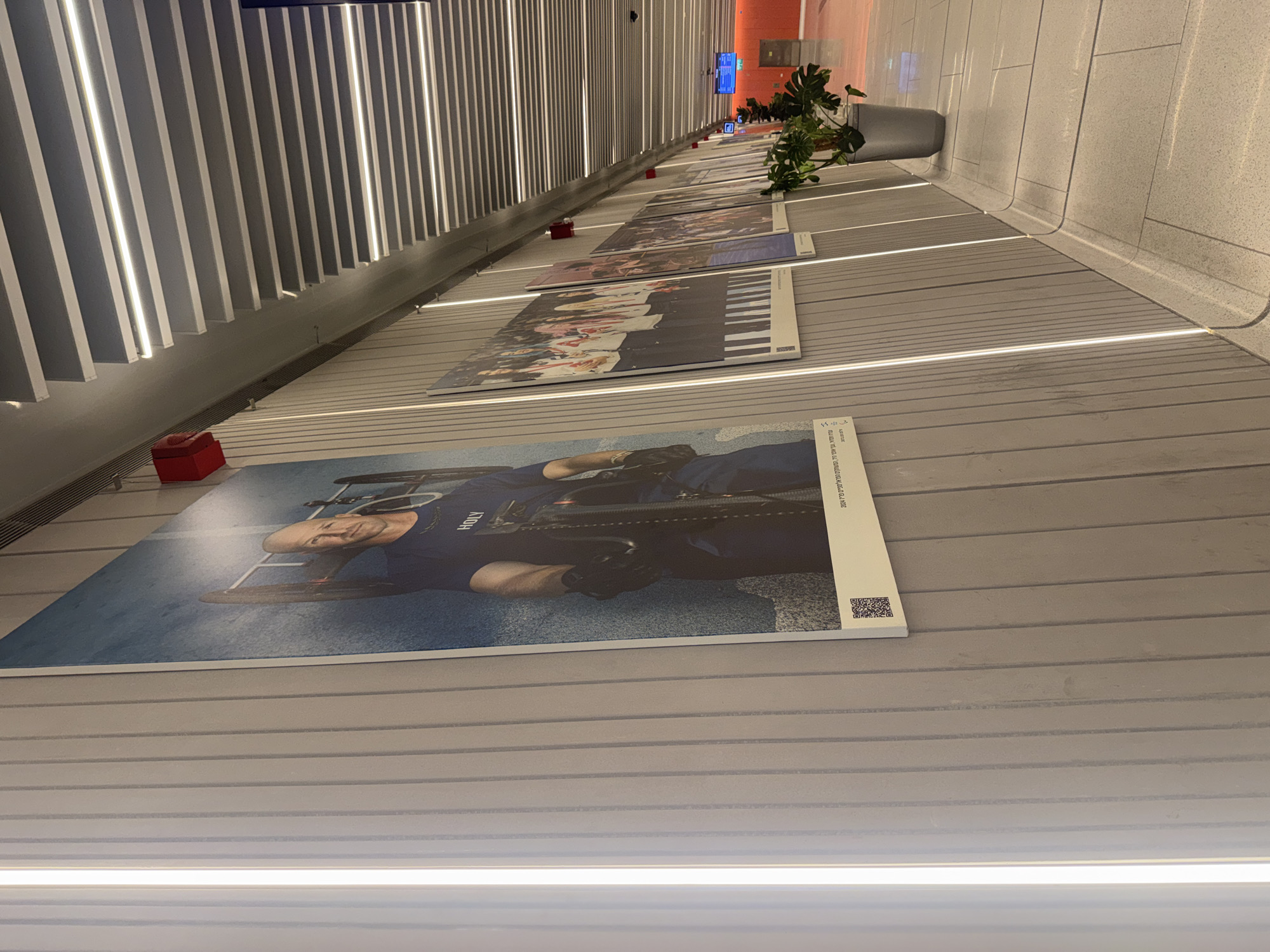 The image shows a series of displayed photographs mounted on a wall, seemingly in an indoor public space in Ramat Gan, Israel. The photographs are arranged linearly along the wall, becoming smaller in perspective towards the vanishing point in the distance. Starting closest to the viewer, the first photograph features a male athlete in sporting attire, possibly a cyclist, with the name "HOLY" visible on his uniform. Subsequent photos depict groups of people and individuals in different settings. A red, square-shaped object sits near the edge of the wall, next to the first photograph. Along the opposite wall, vertical slats with integrated linear lighting illuminate the space. In the distance, some potted plants are visible near the end of the displayed photographs. The floor is light-colored and appears to have a textured surface.