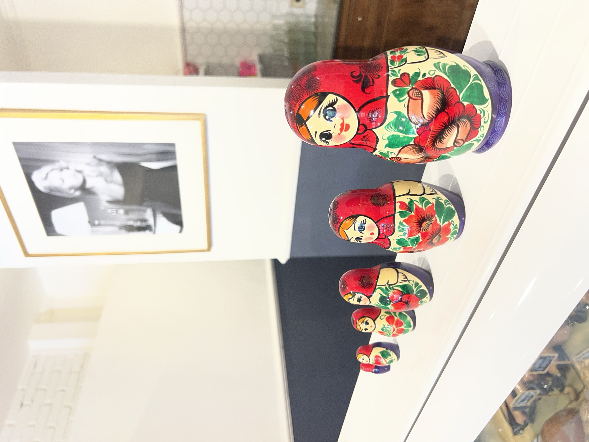 The image shows a set of five Russian nesting dolls (matryoshka dolls) arranged in descending size order on a white shelf or countertop. The dolls are painted with red, yellow, green, and purple hues. All of them depict faces with blue eyes. Behind the dolls, a framed black-and-white photograph hangs on a white wall, with a grey section visible between the wall and shelf. In the background, there are hints of a white-tiled wall and a wooden cabinet. The location of the picture is in North Kuta, Indonesia.