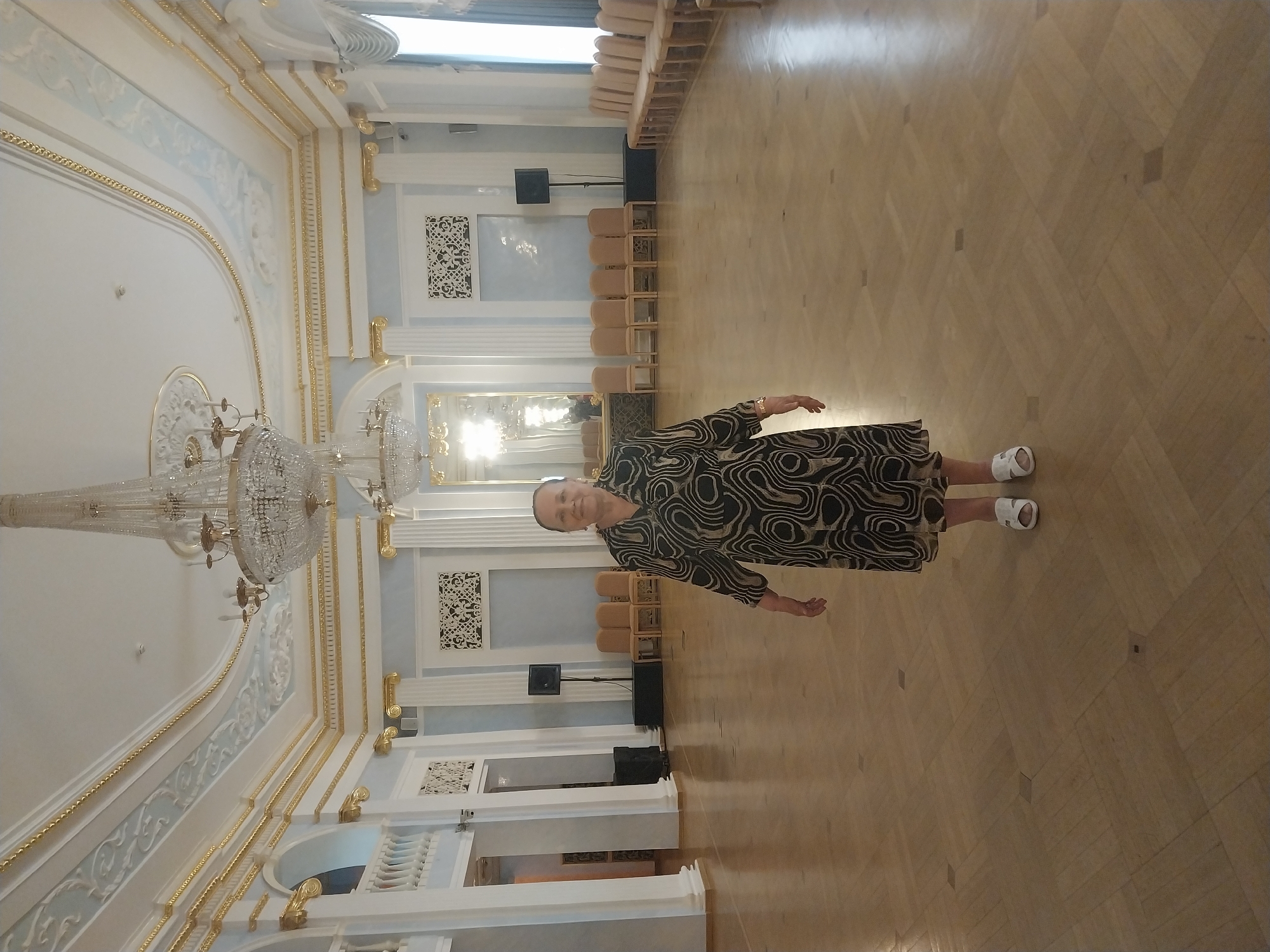 A full shot shows a woman standing in a large, ornately decorated hall. The hall features a polished wooden floor laid in a parquet pattern, high ceilings with intricate white and gold molding, and a large crystal chandelier hanging from the center. The walls are painted a pale blue and adorned with decorative panels and gold trim. In the background, rows of chairs are visible, and sound equipment is set up, suggesting a possible event or performance. The woman is wearing a black and white patterned dress and white slip-on shoes. She is facing the camera with a neutral expression and her right arm extended slightly. The lighting suggests daytime, and the setting appears to be an indoor public space, possibly a ballroom or event hall, consistent with a location in Orenburg, Russia. There is no visible text in the image.