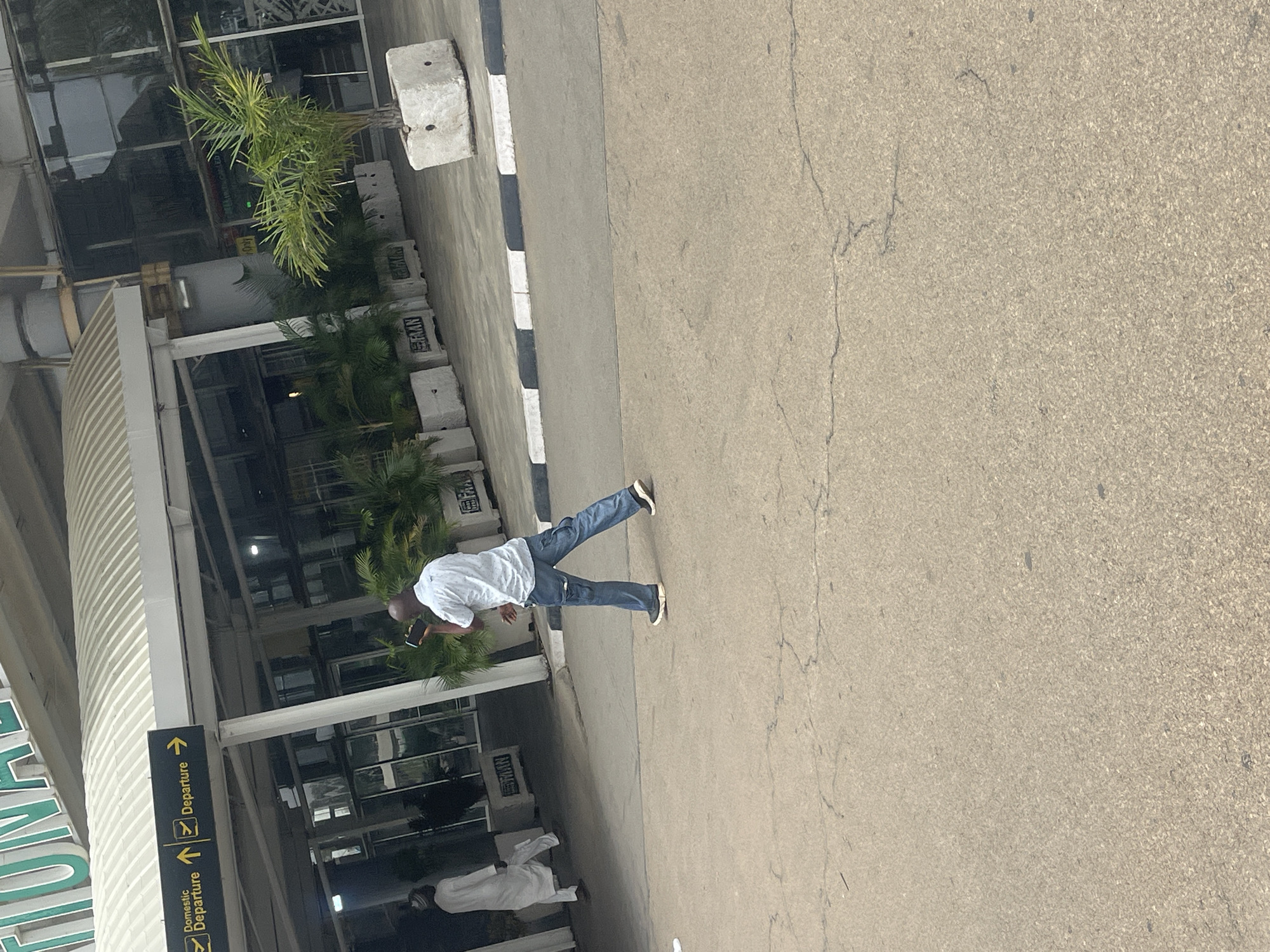 The image, captured in Gwaska, Nigeria, presents an outdoor scene in front of an airport terminal under bright daylight. The main subject is a man wearing a white collared shirt, blue jeans, and dark shoes, holding a smartphone. He is walking on a paved area, seemingly a pedestrian path, with the airport building to his left.
Further back, closer to the building, another person in white traditional attire is visible. The modern airport terminal features extensive glass facades and a distinctive curved, ribbed canopy. Lush green palm trees and other tropical plants are positioned in front of the building, enhancing the landscaping.
A black and white striped curb delineates the paved area from an asphalt road, which shows minor cracks. Along this edge, several concrete barriers are placed, some bearing partially legible text that appears to be "SAAN" or "SARAN."
Prominent directional signs are visible near the building, indicating routes. These black signs with yellow text and arrows read "Departure" multiple times, and one specifically states "Domestic Departure." A large green sign, partially visible on the building's facade, likely spells out "INTERNATIONAL" or the airport's name. The scene suggests a clear, sunny day at a functional airport.