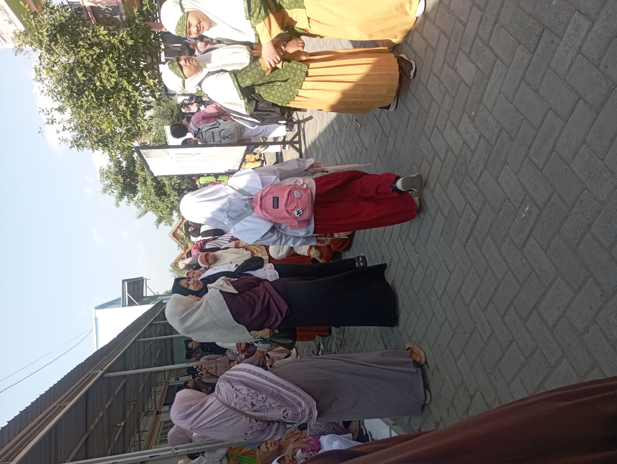 The image depicts a group of people, predominantly women and girls wearing hijabs, gathered outdoors in what appears to be a school or community event in Nganjuk, Indonesia. The setting is paved with interlocking bricks, and there is a covered walkway with a corrugated metal roof to the left, suggesting an outdoor covered area. Trees with green foliage are visible in the background, along with parts of a building. Several individuals are standing and interacting. One young girl in the foreground is wearing a pink backpack and a red skirt, facing away from the camera. Other individuals are dressed in traditional Indonesian attire. The overall atmosphere seems to be one of a social gathering or educational activity.