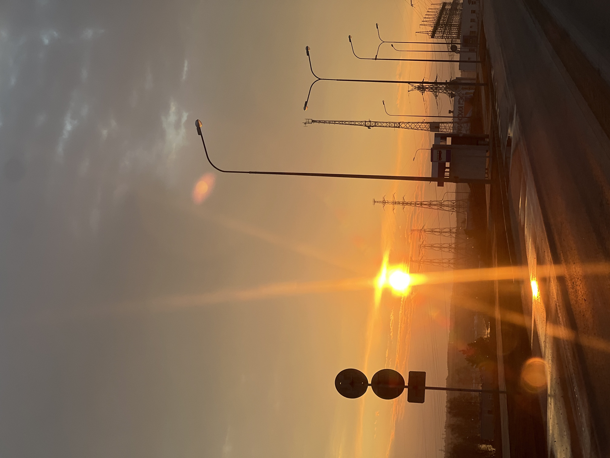 The image shows a sunset in Chaikovsky, Russia. The sun is low in the sky, creating a strong glare. In the foreground is a sign. Behind the sign are power lines and lampposts. The road is to the right. The sky is mostly cloudy.