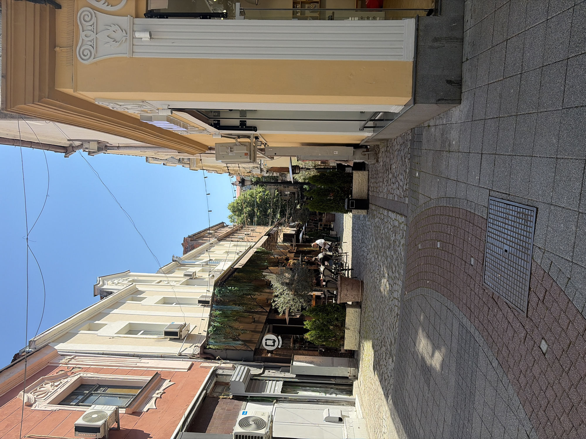 A street in Plovdiv, Bulgaria is visible, looking down a narrow pedestrian thoroughfare. Buildings line both sides, exhibiting architectural details and air conditioning units. Outdoor seating and greenery mark a cafe along the street. Pedestrians are visible at the cafe. The street is paved with a combination of gray and reddish-brown stones, and a manhole cover is also visible. The sky is clear and blue. Overhead wires crisscross the view.