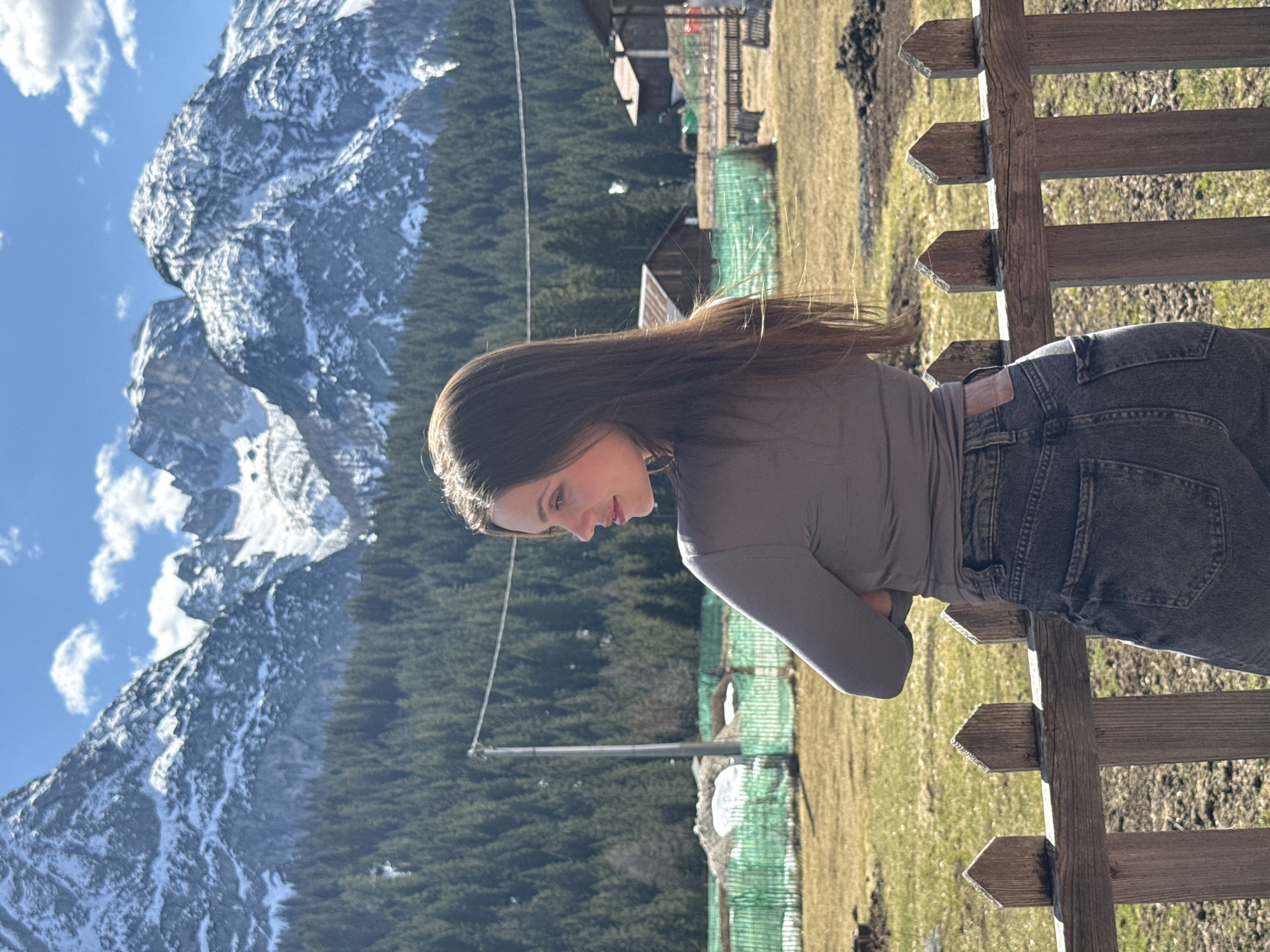 The image depicts a young woman outdoors, leaning against a wooden fence. She is wearing a long-sleeved gray shirt and dark-wash jeans. The background showcases a mountainous landscape, partially covered in snow, likely the Dolomites. A dense forest extends toward the mountains. A building is visible in the distance. The setting appears to be in or near Sappada/Plodn/Sapade, Italy, based on the characteristic mountain scenery. The time of day appears to be daytime with ample sunlight.