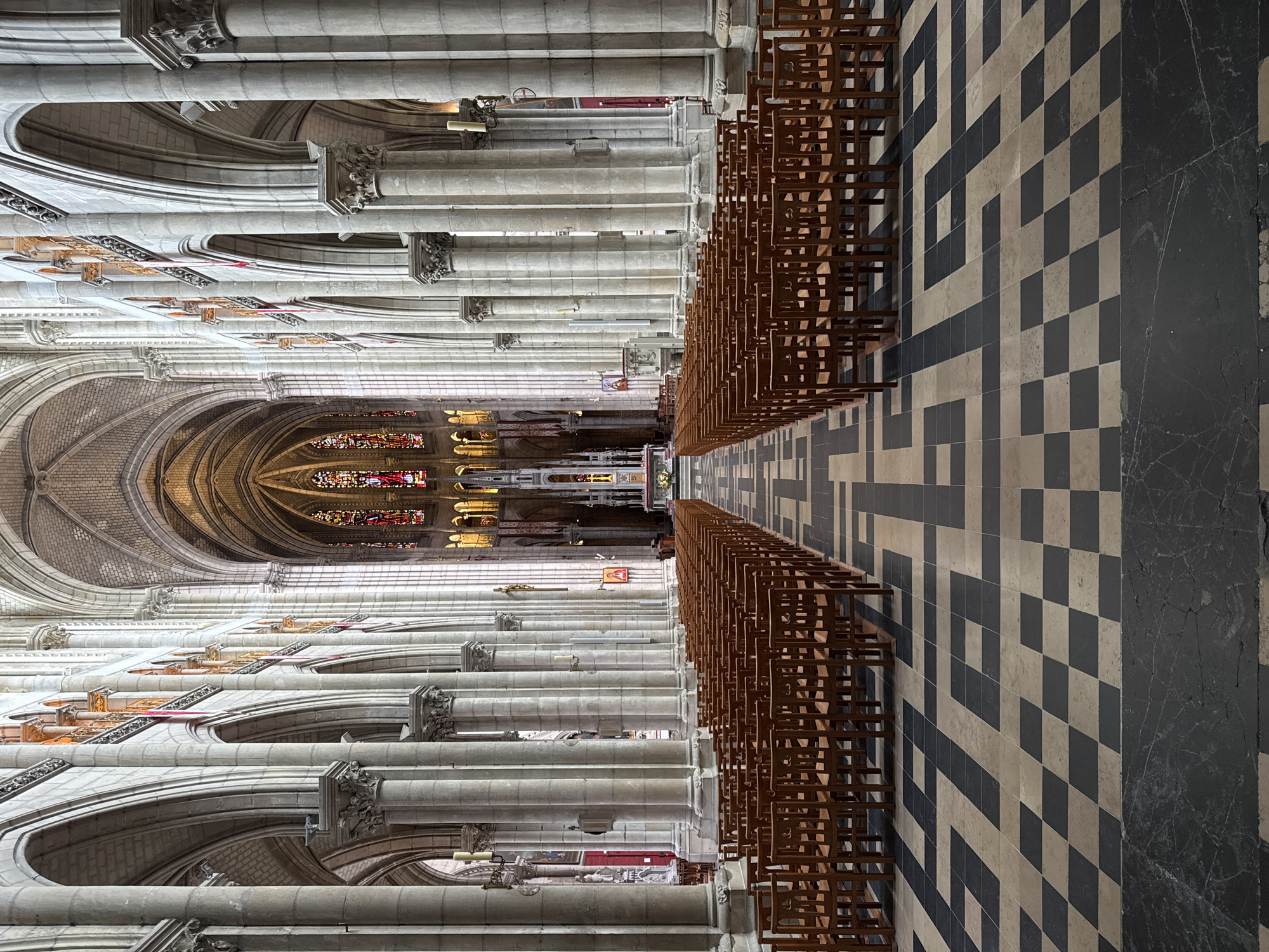 The image shows the interior of a large Gothic cathedral, likely the Nantes Cathedral in Nantes, France, based on the architectural style and context. The perspective is from the central aisle, looking towards the altar at the far end. Rows of empty wooden chairs are arranged on either side of the aisle. The floor is tiled in a distinctive pattern of alternating light and dark squares and rectangles. Massive stone columns with ornate capitals line the nave, supporting arched ceilings. Stained-glass windows with colorful religious imagery are visible along the upper walls. The overall lighting is dim, with light filtering through the windows and from unseen sources, creating a solemn and grand atmosphere. There are no people visible in the image.