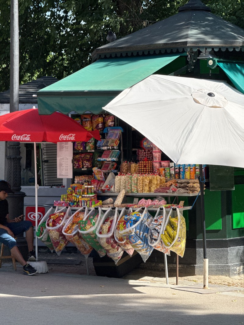 An outdoor snack kiosk is captured on a sunny day in what appears to be a park or public square. The kiosk features a dark green, multi-tiered roof and green awnings, suggesting a permanent or semi-permanent structure. A pigeon is perched on the peak of the kiosk's roof.
In the foreground, a large white umbrella provides shade over a section of the kiosk, partially obscuring the view of the items behind it. To the left, a smaller red umbrella with "Coca-Cola" written in white script provides shade for another part of the setup. Underneath this red umbrella, a display of bottled drinks and possibly an ice cream freezer is visible, marked with the red "Iglú" or "Igloo" logo.
The kiosk is heavily stocked with a variety of packaged snacks and candies. Rows of chips, sweets, and other treats are displayed on shelves and hanging racks. Prominently featured are numerous clear plastic bags, each filled with brightly colored snacks, such as popcorn, chips (including visible bags of "Doritos" and "Ruffles"), and other savory treats. These bags are suspended from hooks along the front of the display, creating a colorful and inviting visual.
A young man, possibly a customer or a vendor taking a break, sits on a small wooden stool to the left, under the red Coca-Cola umbrella. He is wearing a dark t-shirt, blue shorts, and a baseball cap, and appears to be looking at his mobile phone. His feet, clad in sneakers, rest on the ground. Shadows from the umbrellas and surrounding trees are cast across the paved ground, indicating bright daylight. The overall scene suggests a casual vending point offering refreshments and snacks.