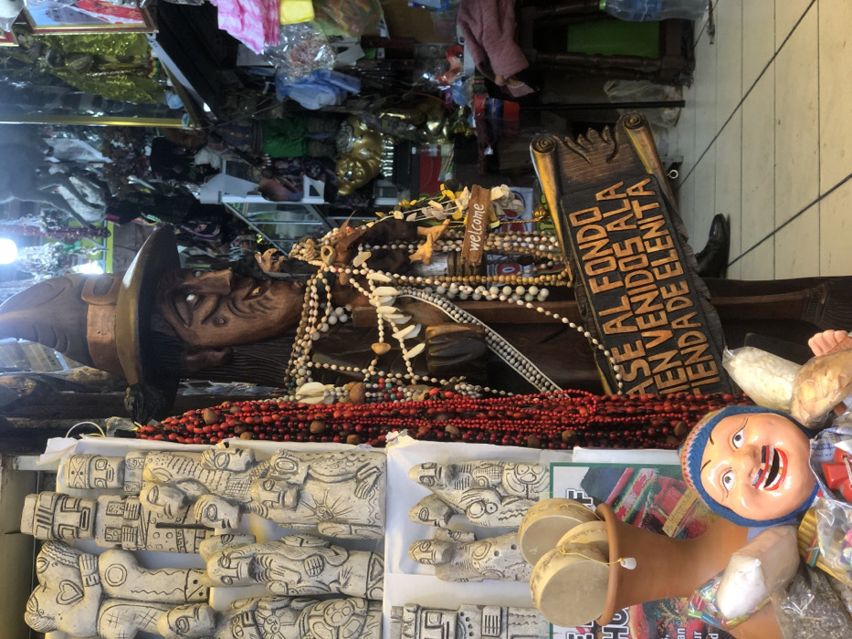 A crowded marketplace scene showcases an array of cultural items. Prominently, a wooden statue of a person with a hat is adorned with beads and shells. Red beads cascade over a display of carved stone figures. A painted mask with a blue hat smiles beside musical instruments. A wooden sign with text is visible, possibly indicating a shop's name. The background reveals an abundance of merchandise.