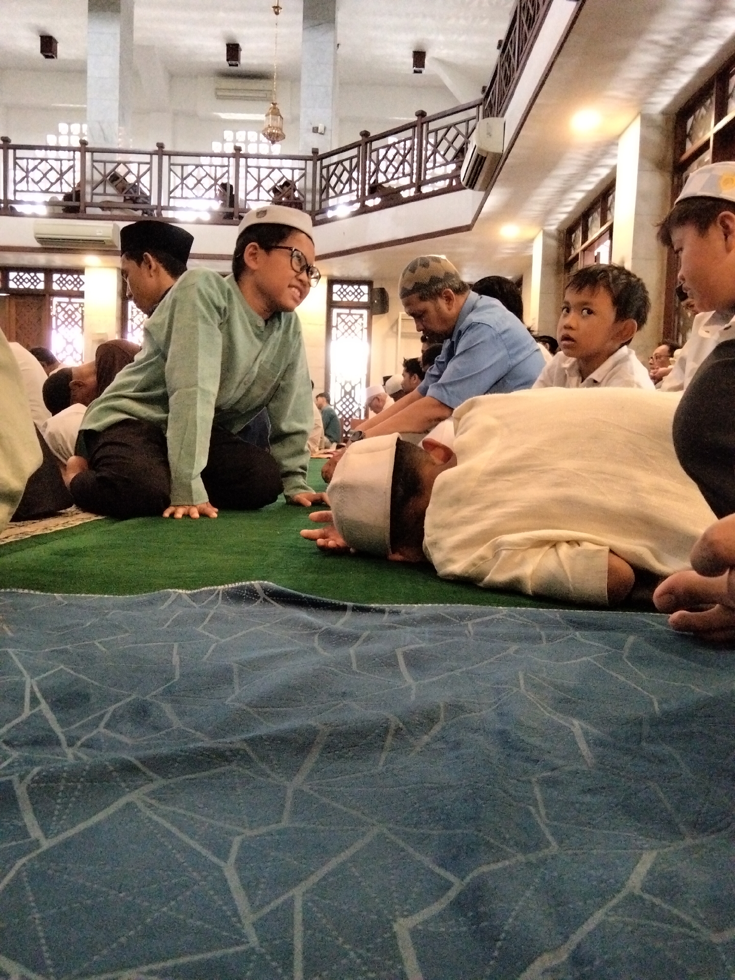 The image depicts a group of people, predominantly young boys and men, inside a mosque, possibly during prayer or a religious gathering. They are dressed in traditional Islamic attire, including skullcaps and long shirts. The mosque's interior is visible, with architectural features like wooden railings and latticed windows. The flooring is covered with green carpet, and a patterned blue rug is in the foreground. The lighting suggests an indoor environment with natural light filtering in and artificial lights illuminating the space. The presence of people in prayer positions and the mosque setting strongly indicate a religious context. The image is likely taken in Depok, Indonesia, a city known for its significant Muslim population and numerous mosques.