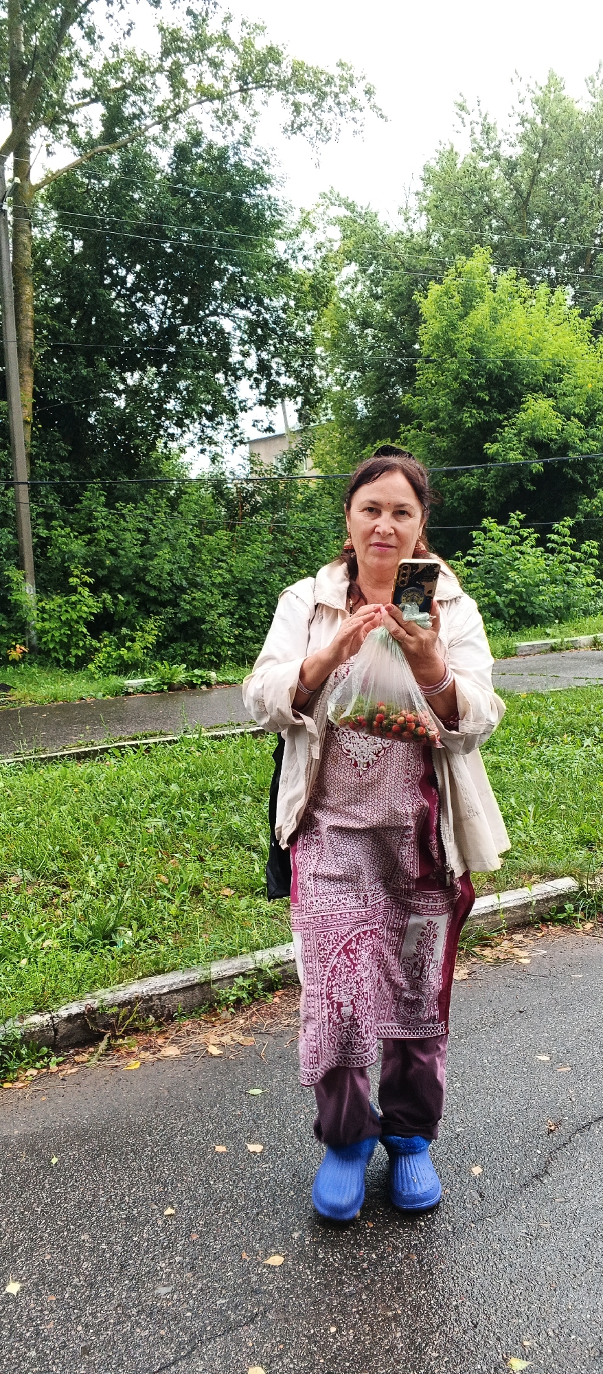 A woman stands outdoors in a paved area, possibly a road or sidewalk, in the city of Podgornoye, Russia. She is holding a clear plastic bag filled with what appears to be small red fruits, likely strawberries or cherries. In her right hand, she is holding a smartphone, which she is looking at. She is wearing a light-colored jacket over a long, patterned tunic and dark pants. On her feet, she is wearing bright blue rubber boots. The background is dominated by lush green trees and foliage, suggesting a natural or park-like setting within the city. The ground is wet, indicating it may have recently rained or is damp. There is a concrete curb and a grassy area separating the paved surface from the vegetation. A utility pole and wires are visible to the left of the woman.