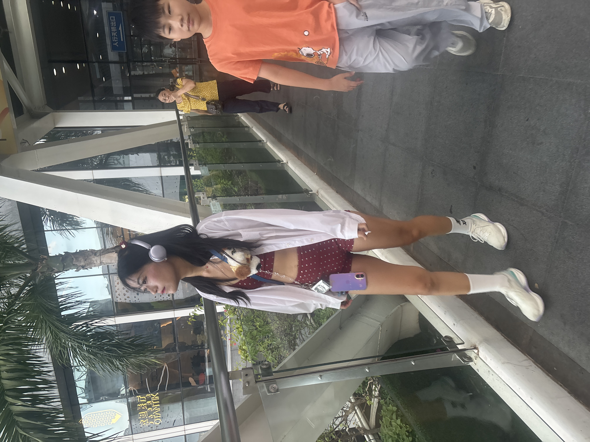 An eye-level, medium shot captures a young Asian woman walking on a pedestrian overpass in the Liwan District of Guangzhou, China. She is wearing a white unbuttoned shirt over a maroon polka-dot bralette and matching shorts, white socks, and white sneakers. She has silver headphones on and is looking to her left. A small plush toy dog is attached to a strap on her chest, and a purple phone case hangs from a blue lanyard around her neck. In the background, to her right, a woman in a yellow polka-dot shirt and dark pants is walking away, and a young Asian boy in an orange t-shirt with a Snoopy graphic and light gray pants is walking towards the camera. The overpass has a glass railing on the left, overlooking lush greenery below. The background features a modern building with large glass windows.