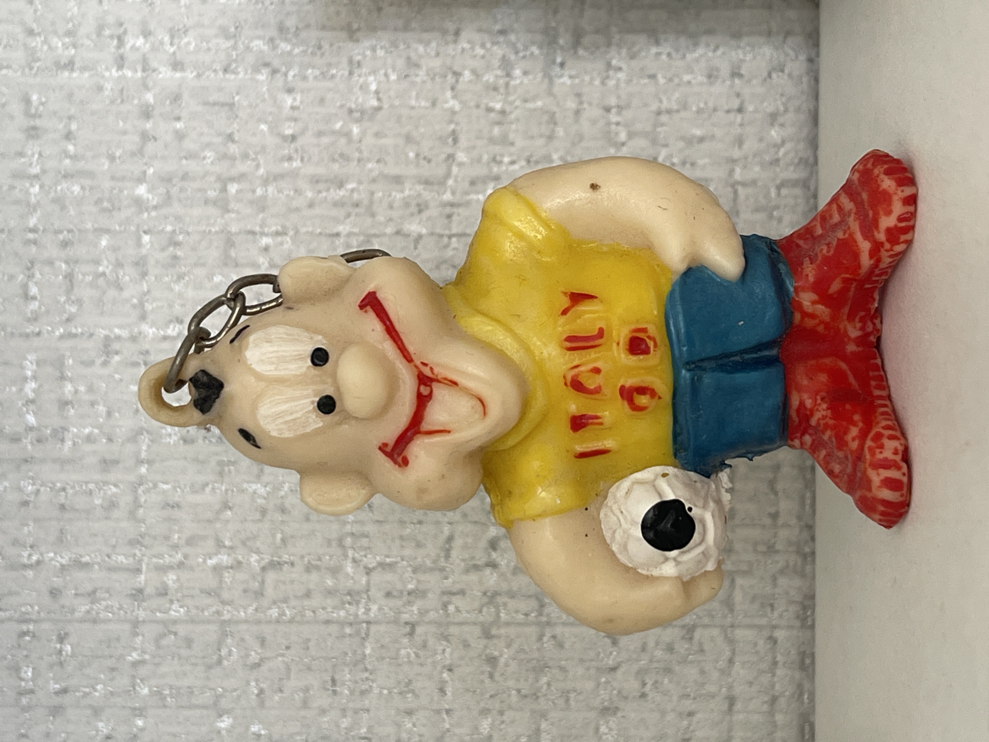 The image shows a keychain figure of a soccer player wearing a yellow shirt that says "ITALY," blue shorts, and red and white patterned boots. The figure is pale, with black dots for eyes and a red design on the face. It is holding a soccer ball. The keychain is made of metal. The background is a textured gray surface. The location in the city of Перевозное, Russia is not discernible from the image.