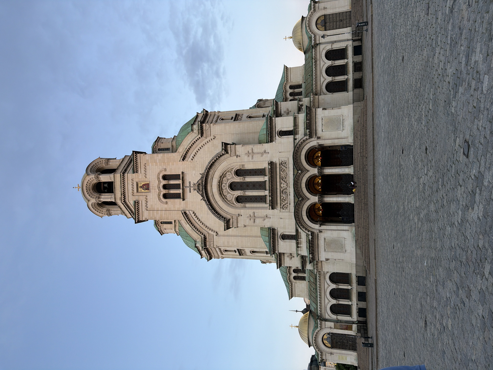 The image shows the Alexander Nevsky Cathedral in Sofia, Bulgaria. The cathedral is a large, ornate structure with a tower and domes. The architectural style is Neo-Byzantine. The building is white or off-white, and the domes have a green tint. The sky is blue with some light clouds. The foreground consists of a cobblestone pavement. No people are readily visible in the image. The camera appears to be tilted on its side.
